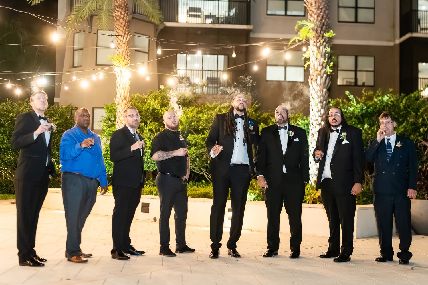 Groomsmen in black suits pose outdoors at night under string lights near palm trees.