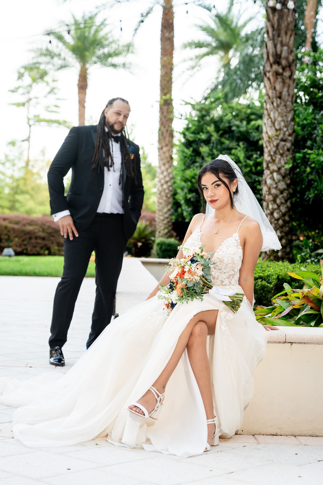 Bride in lace gown seated with bouquet as groom stands behind among palm trees.