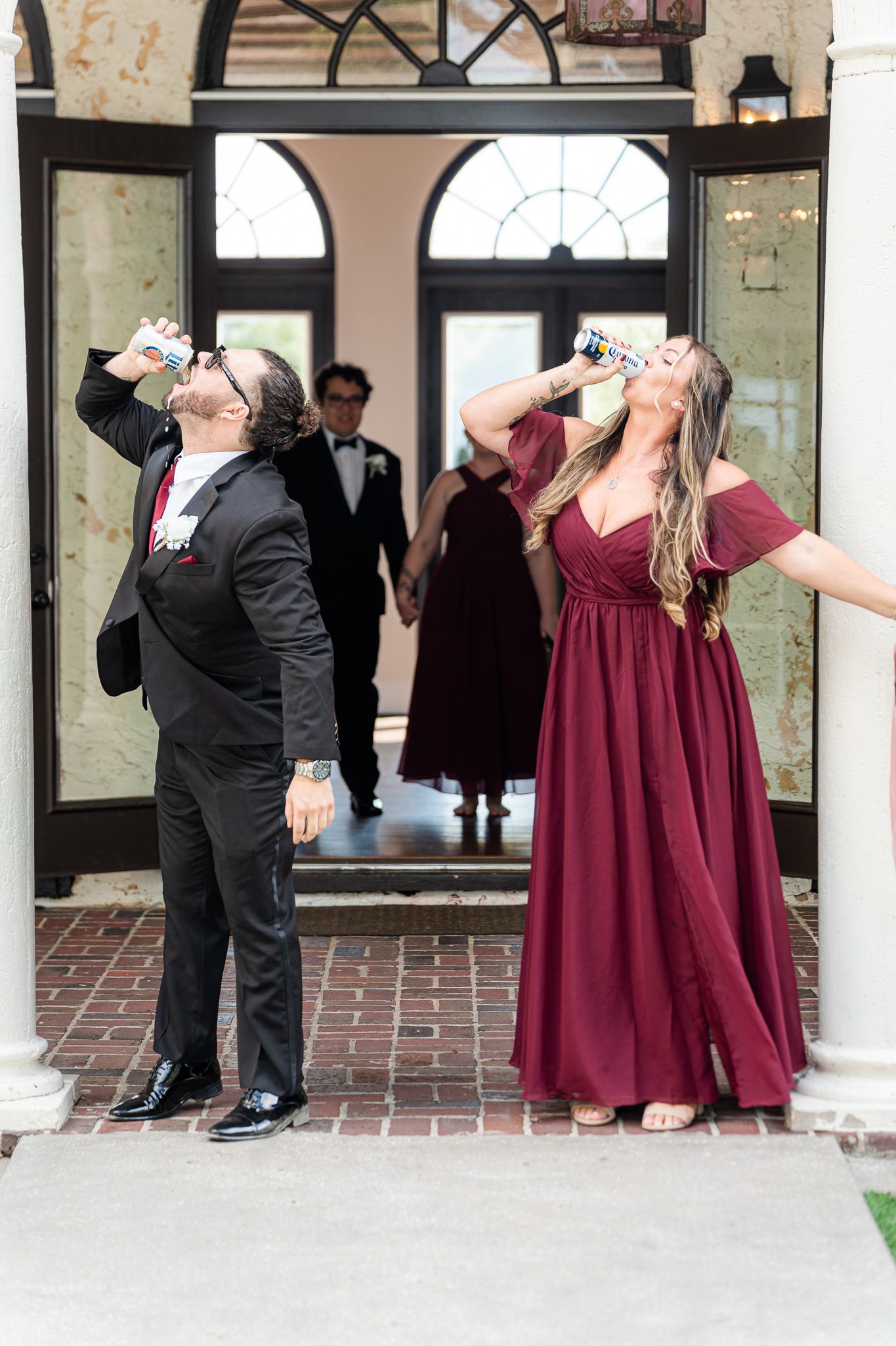 Wedding guests in burgundy and black attire taking celebratory shots outside elegant venue doors.