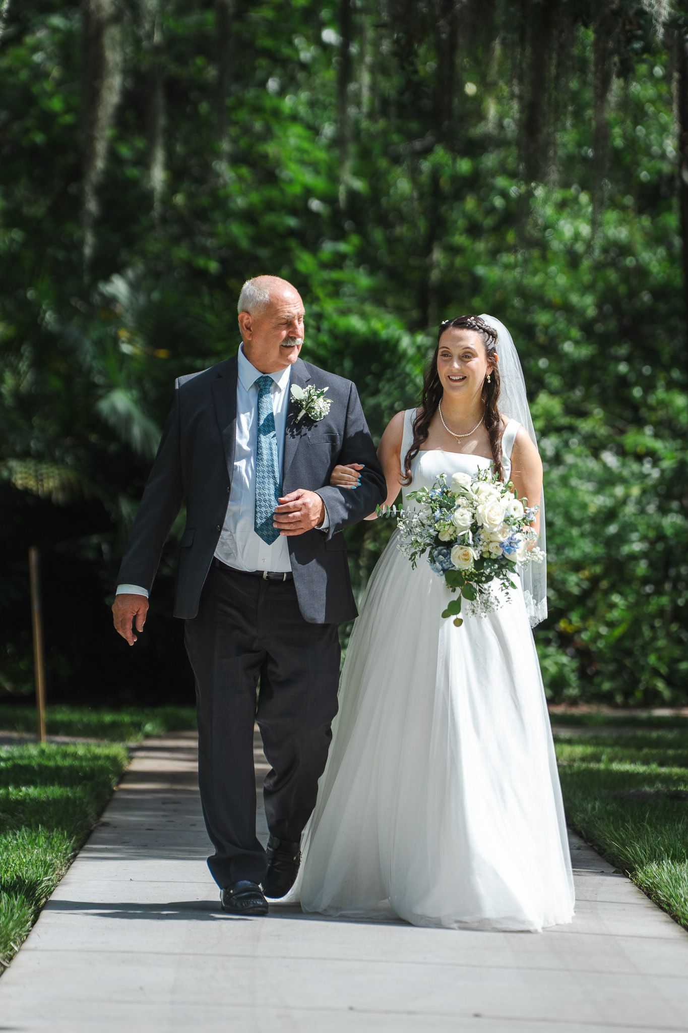Smiling bride in white gown walks down the aisle with her father at an outdoor garden wedding.