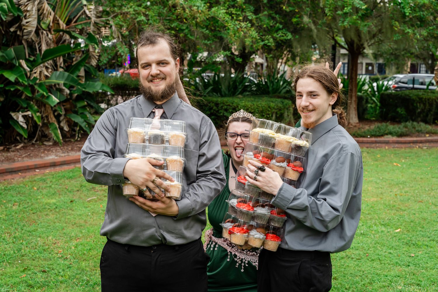 Three smiling staff members in gray uniforms holding clear packaged dessert cups outdoors in a garden setting.