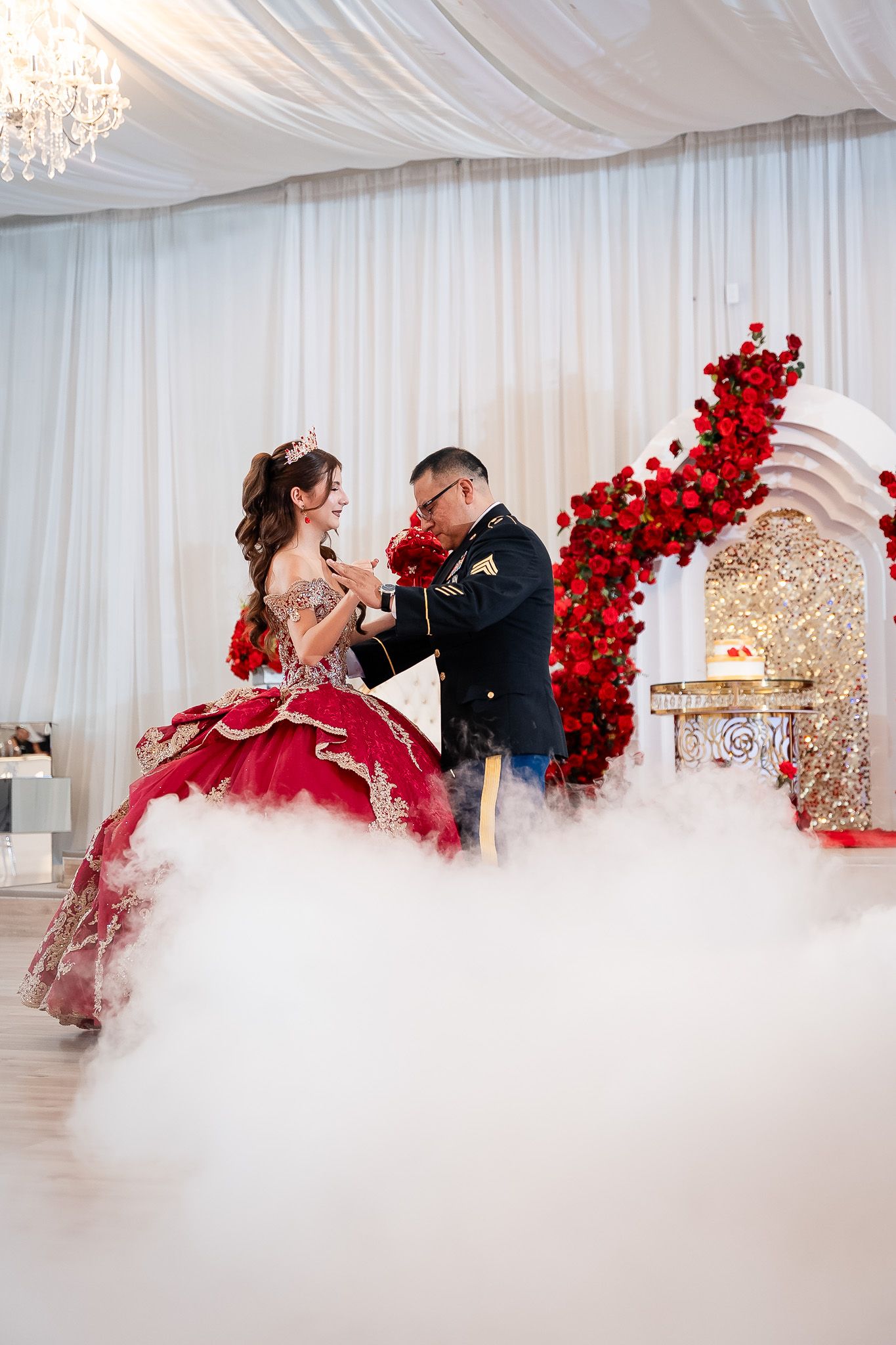 A young woman in a red quinceañera gown dances with a man in military uniform amid dramatic fog effects at an elegant venue.