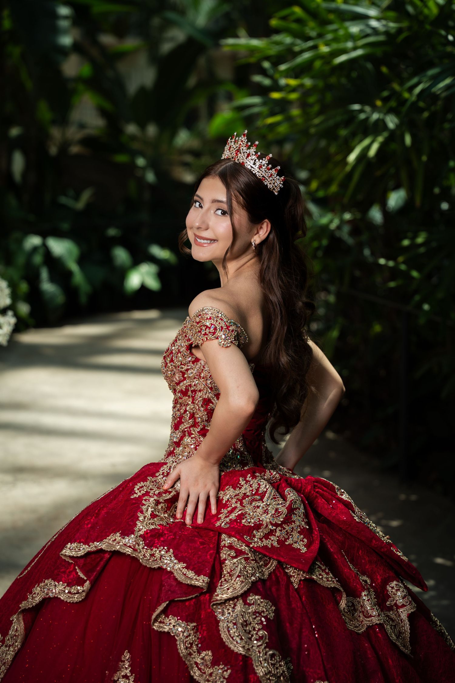 Young woman in red and gold quinceañera ball gown with tiara poses gracefully in lush garden setting.