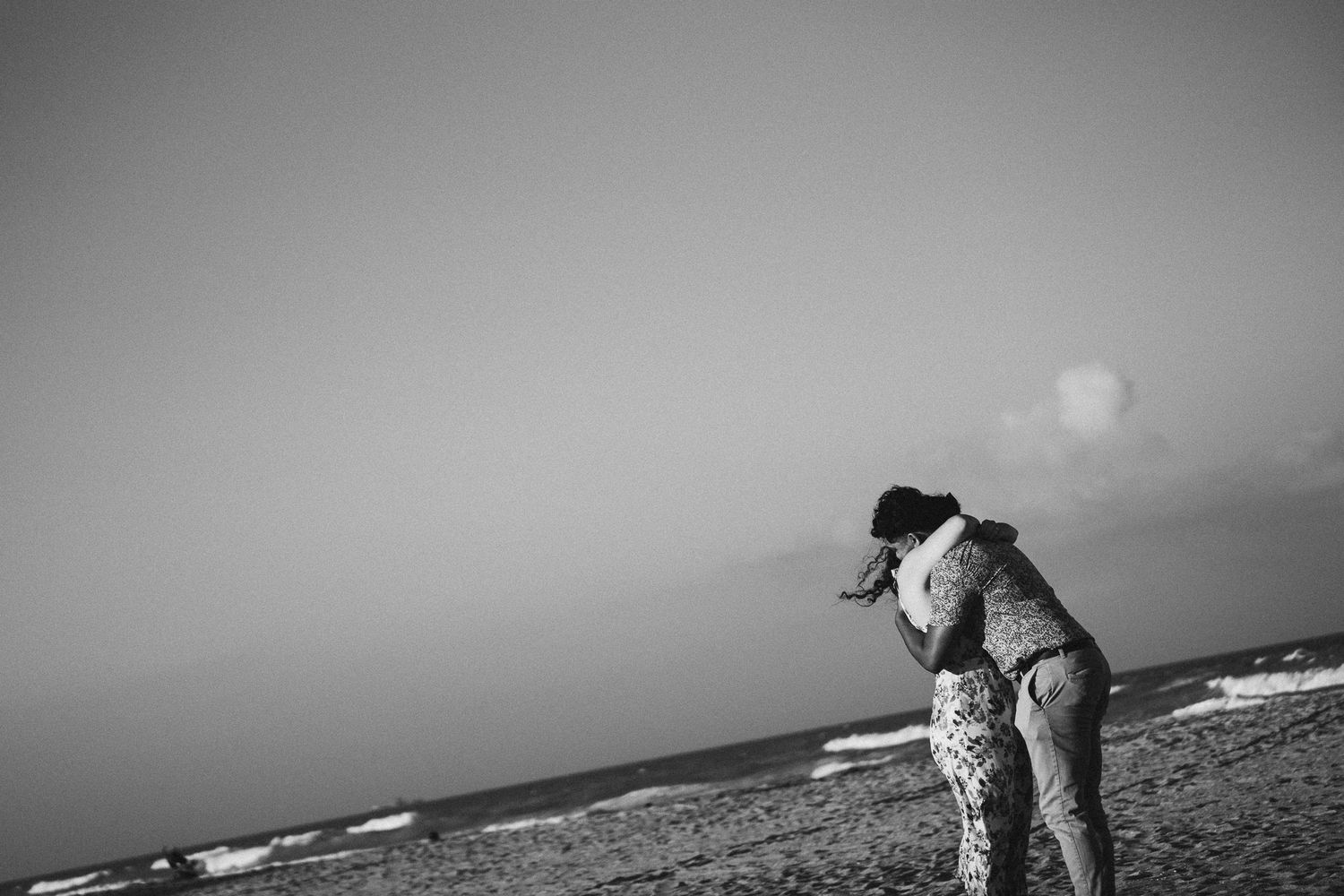 Black and white photo of a couple embracing on a sandy beach with ocean waves in the background.