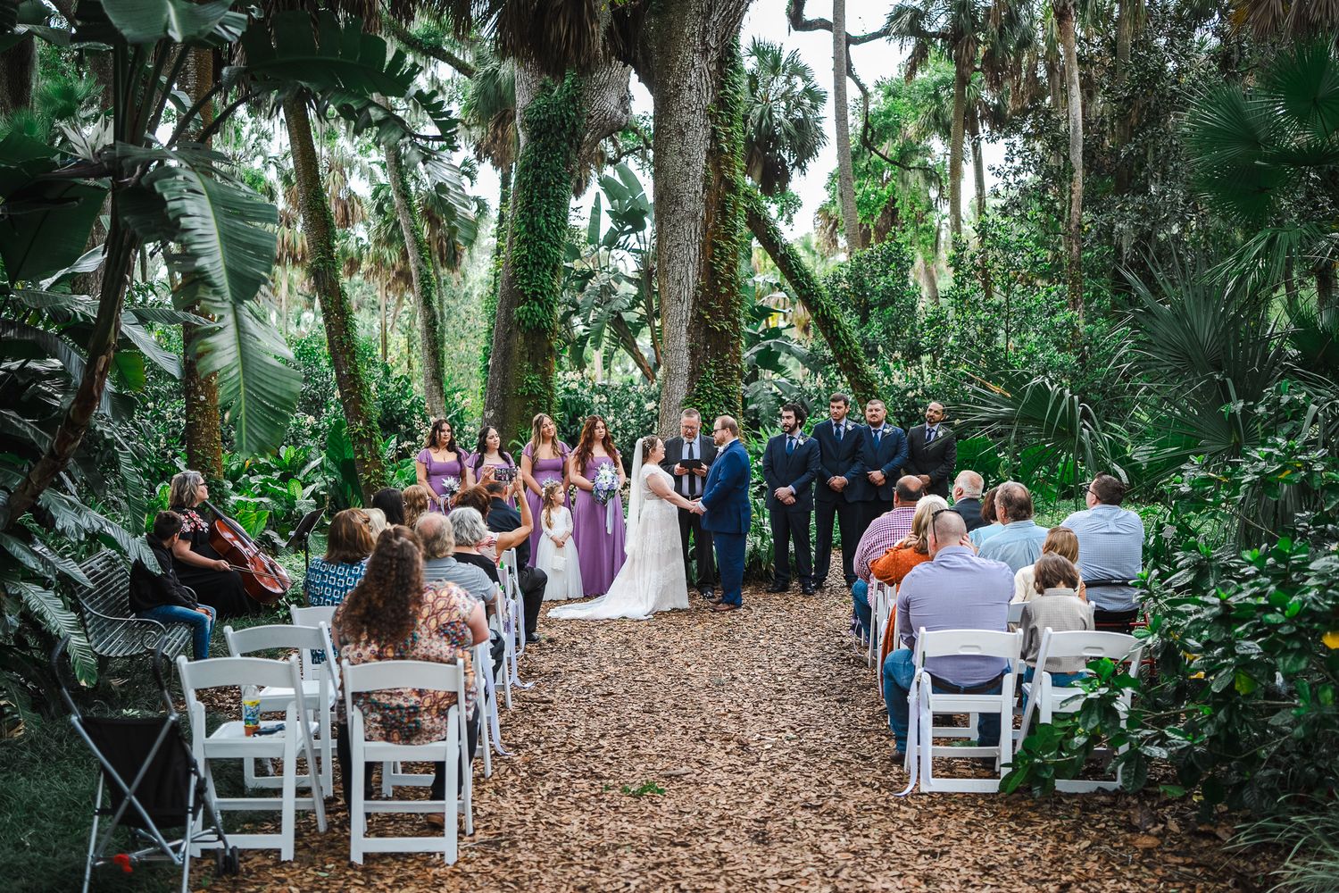 Outdoor wedding ceremony in a lush tropical garden with guests seated in white chairs along a tree-lined aisle.