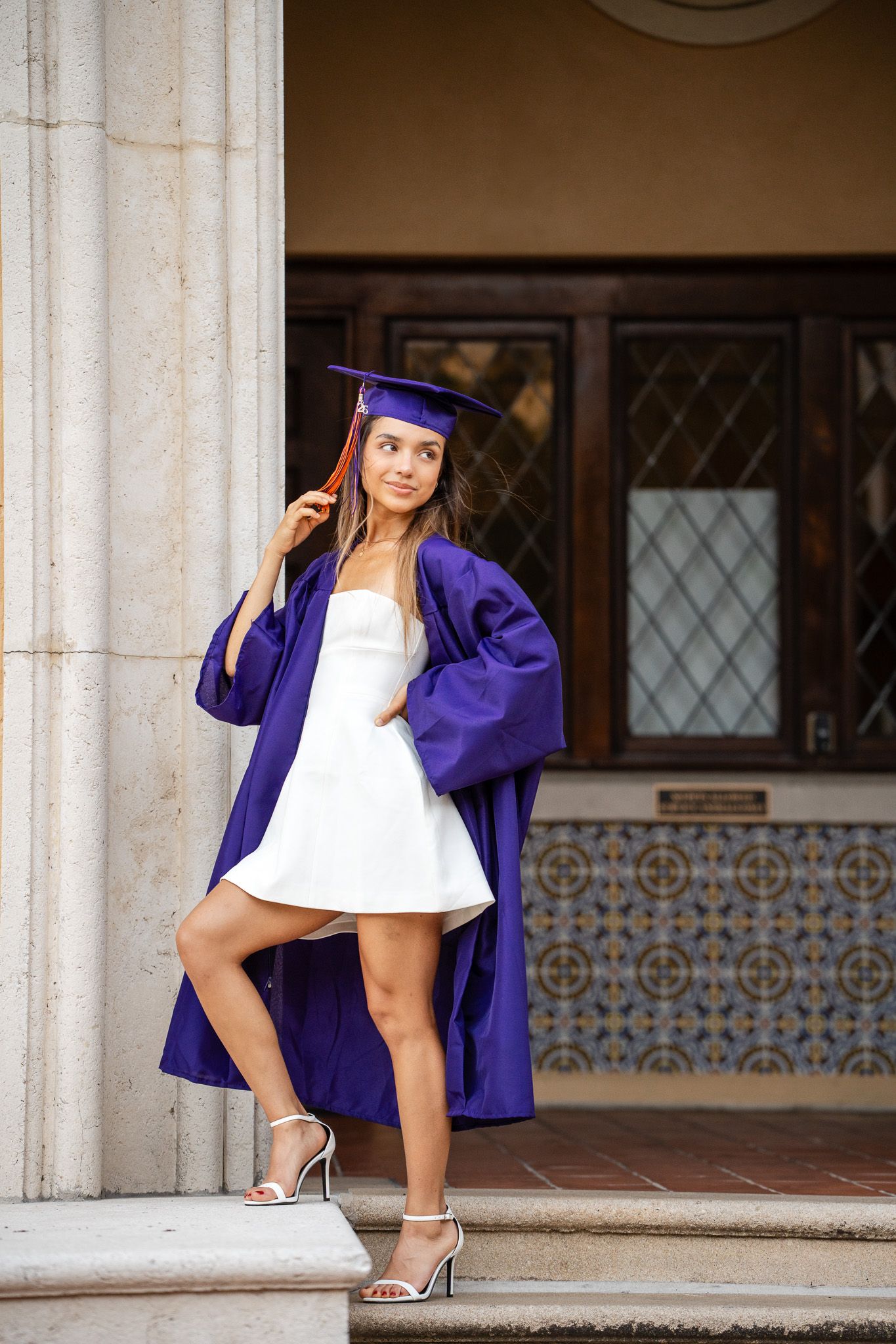 Graduate in purple cap and gown posing confidently by ornate columns of a classic building.