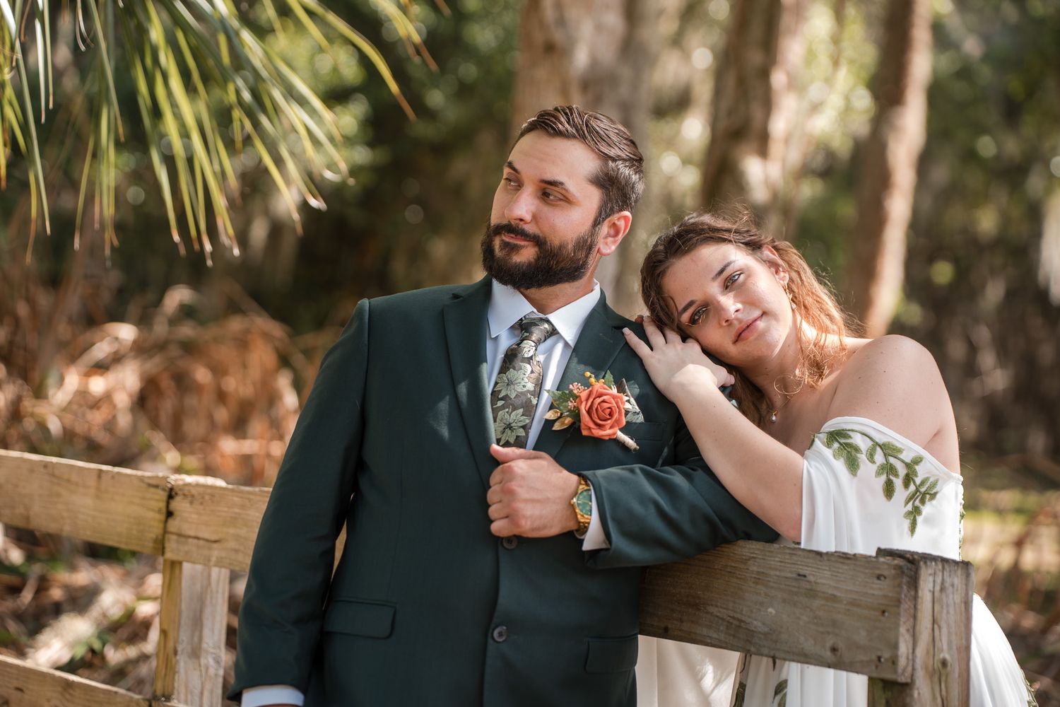 Happy couple poses on wooden bridge, groom in dark suit with floral tie, bride in off-shoulder gown.