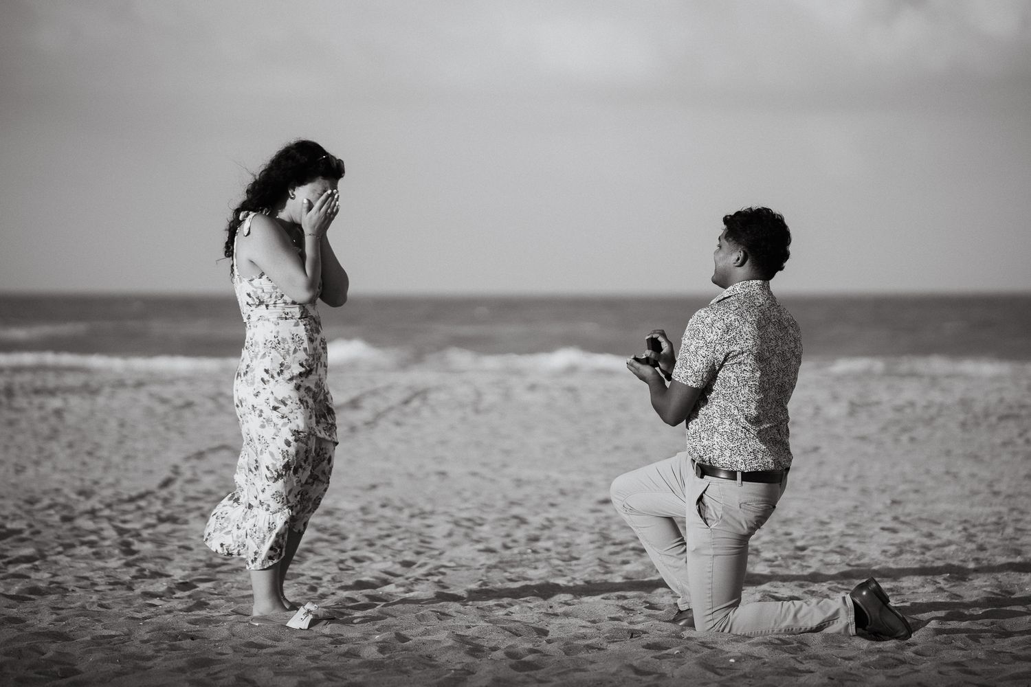Black and white photo of a man on one knee proposing to a woman on the beach.