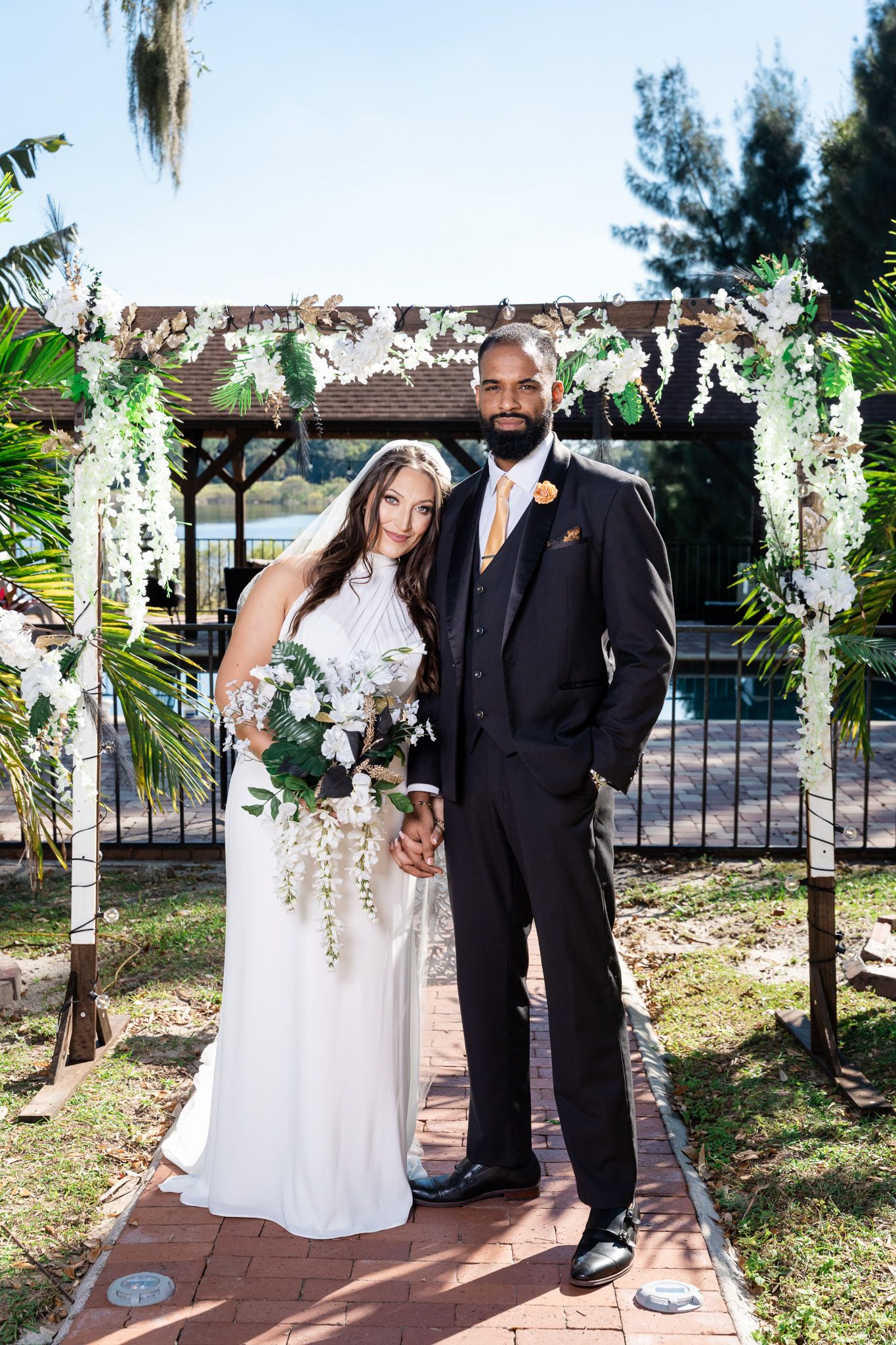 Interracial wedding couple posing under floral arch decorated with white flowers and greenery outdoors.