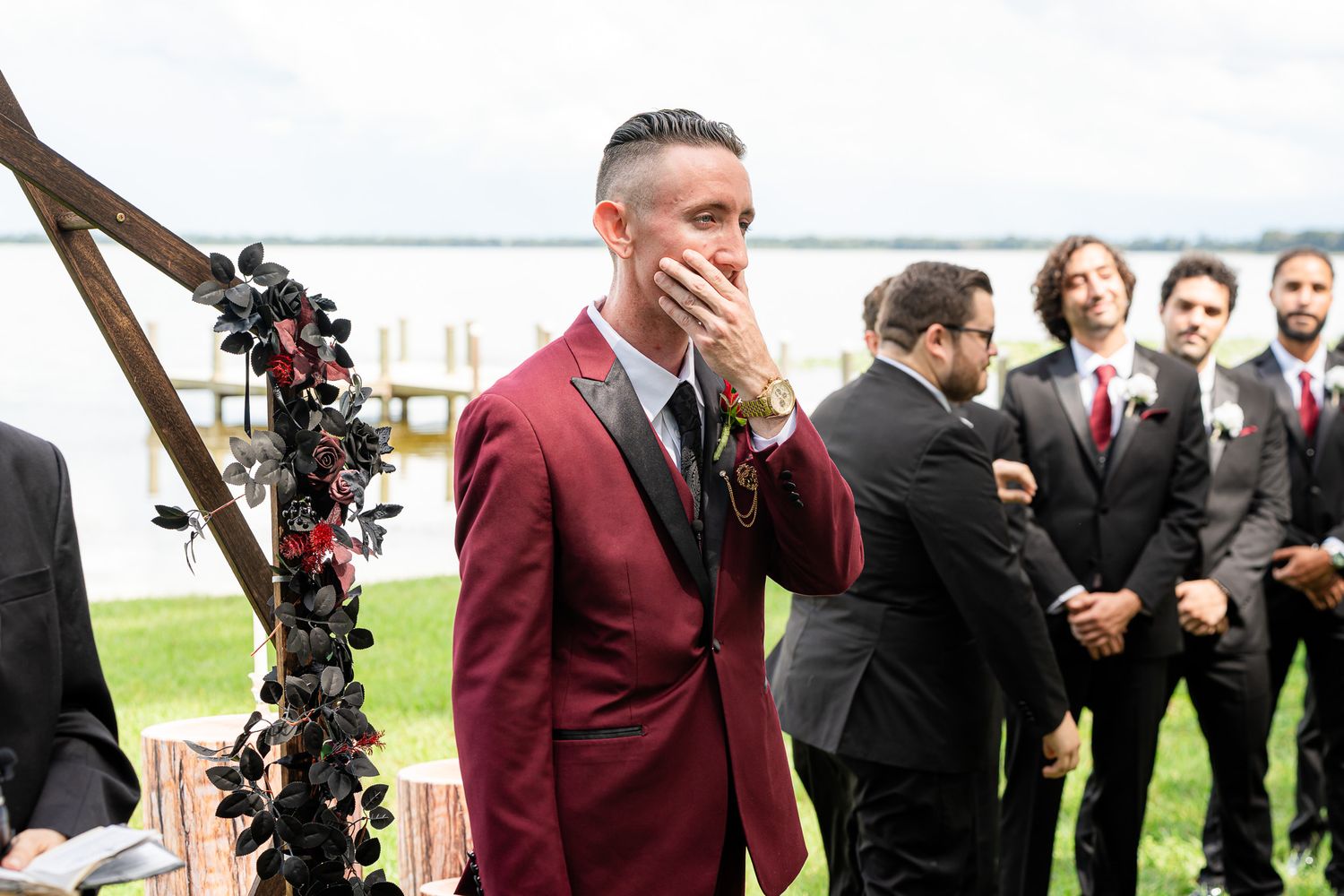 Emotional groom in burgundy suit covers mouth as bride approaches outdoor waterfront ceremony.