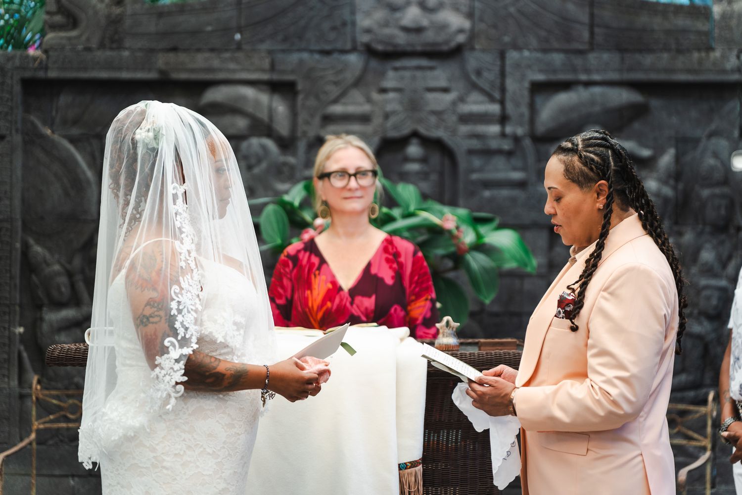 Two brides exchange vows at an outdoor wedding ceremony with an officiant in a red dress.