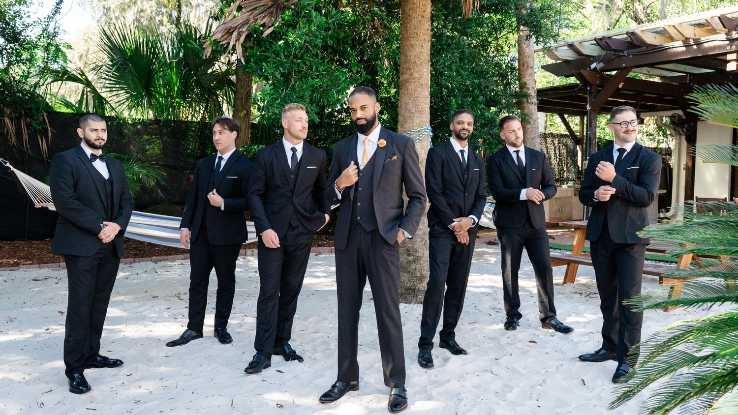 Groom in dark suit stands confidently with groomsmen in black tuxedos on a sandy outdoor area.