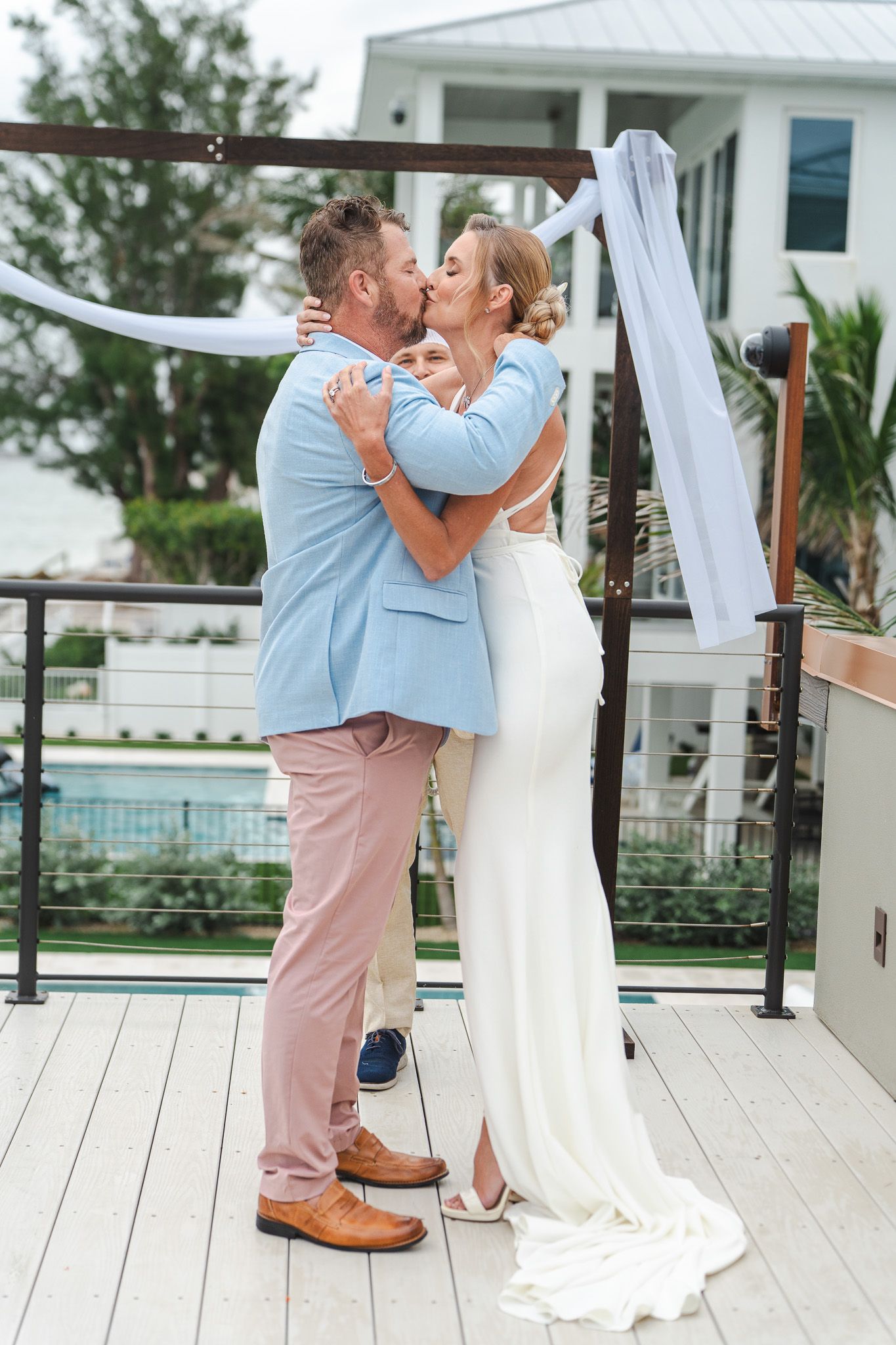Bride and groom share first kiss at outdoor beach wedding ceremony with white fabric arch backdrop.