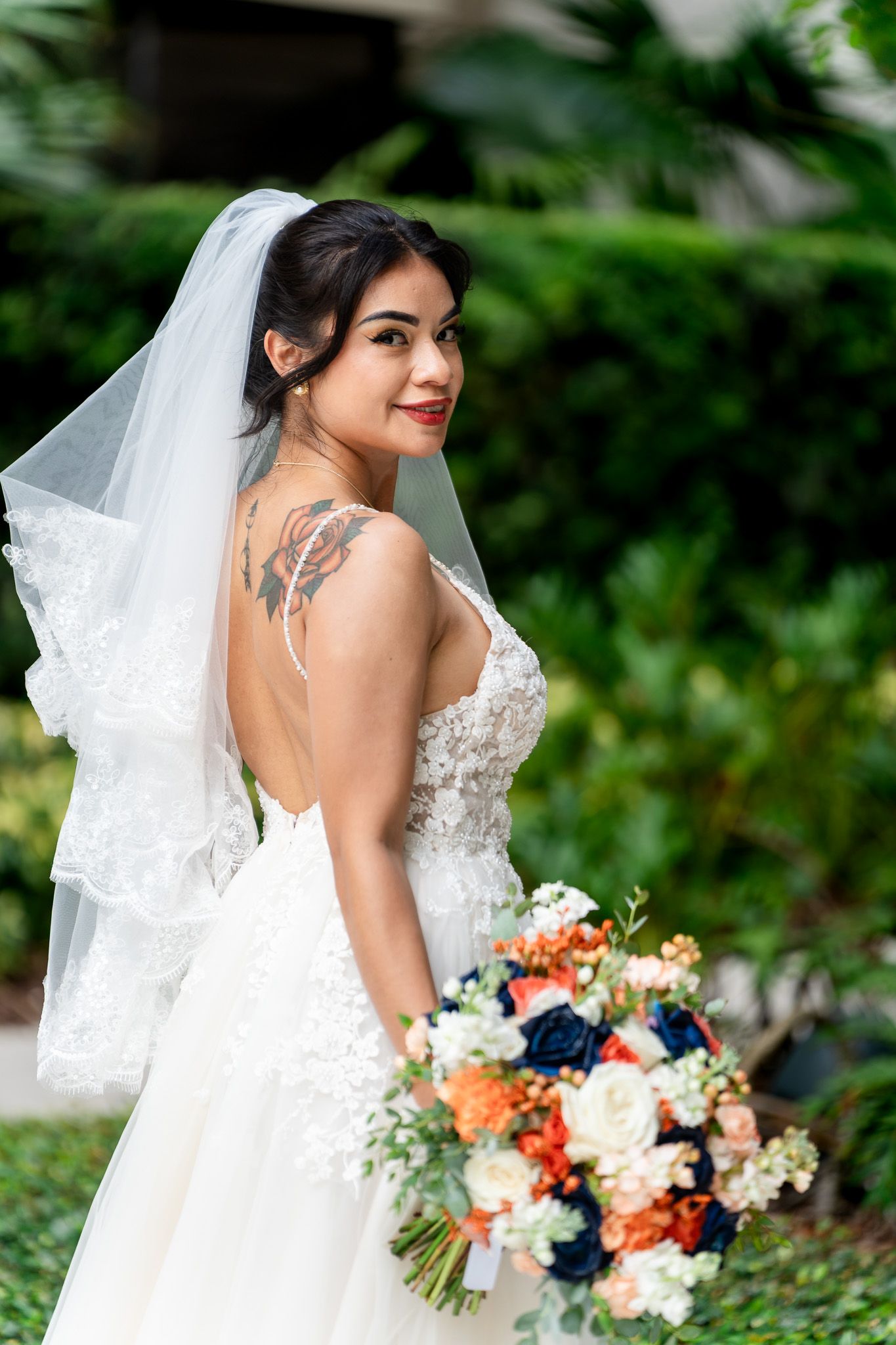 Bride in backless lace gown with floral tattoo holds colorful bouquet of navy and orange roses.