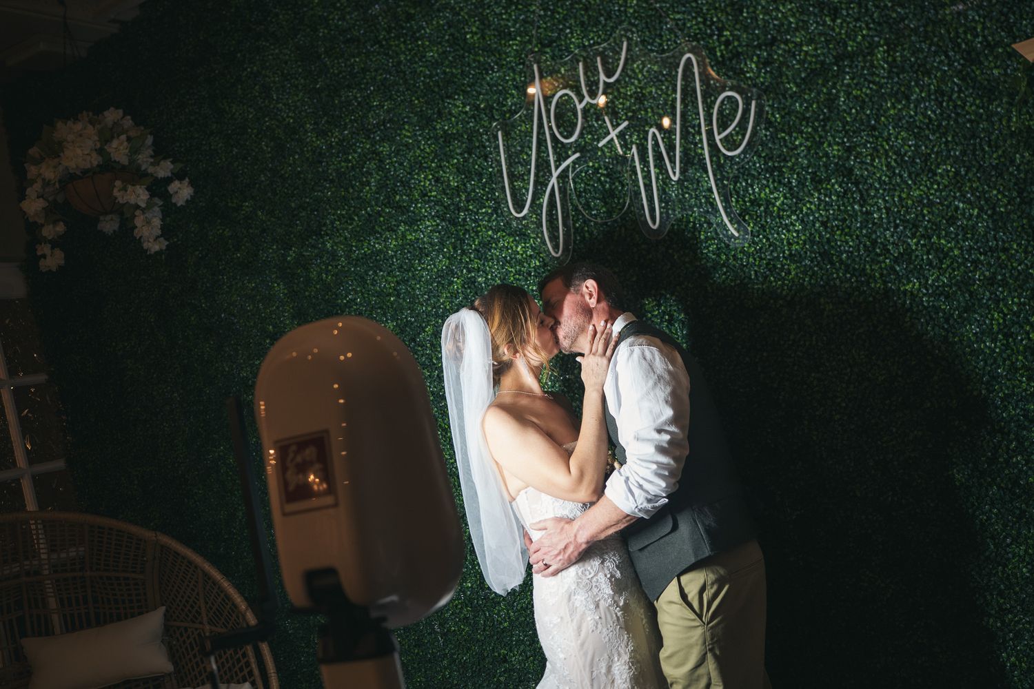 Bride and groom share a romantic kiss in front of a green hedge wall with a glowing 'You & Me' neon sign.