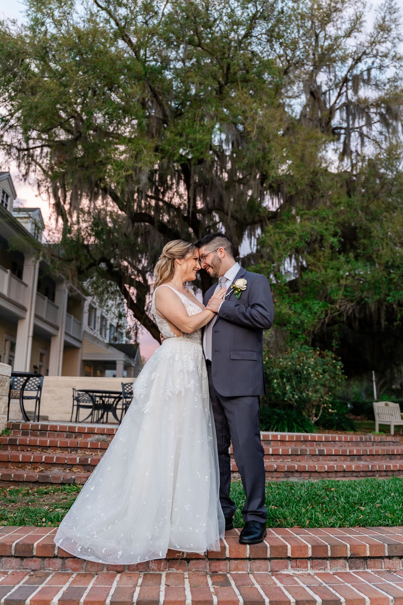 Bride and groom share an intimate forehead touch on brick steps surrounded by lush greenery.
