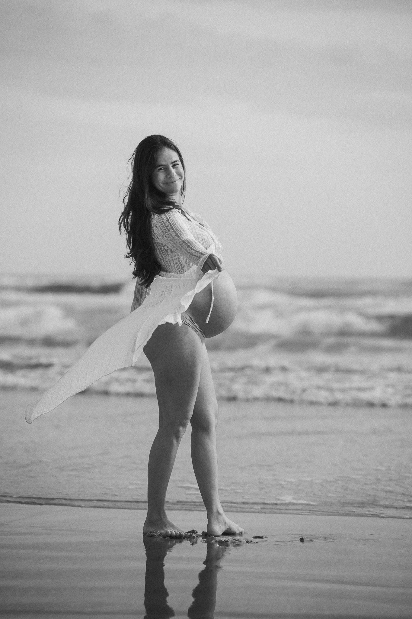 Pregnant woman in white dress posing on beach in black and white maternity photo.