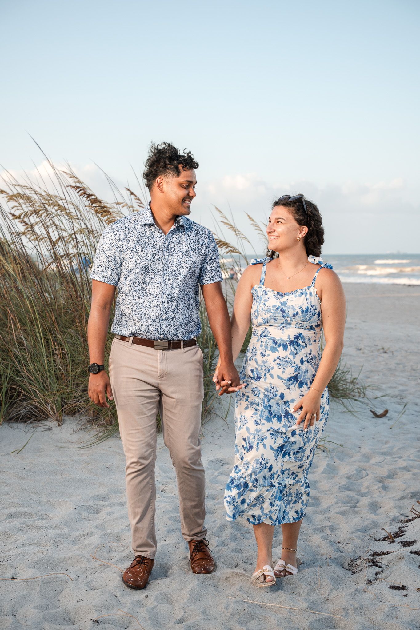 Couple holding hands and smiling at each other on a sandy beach, wearing coordinating blue floral outfits.