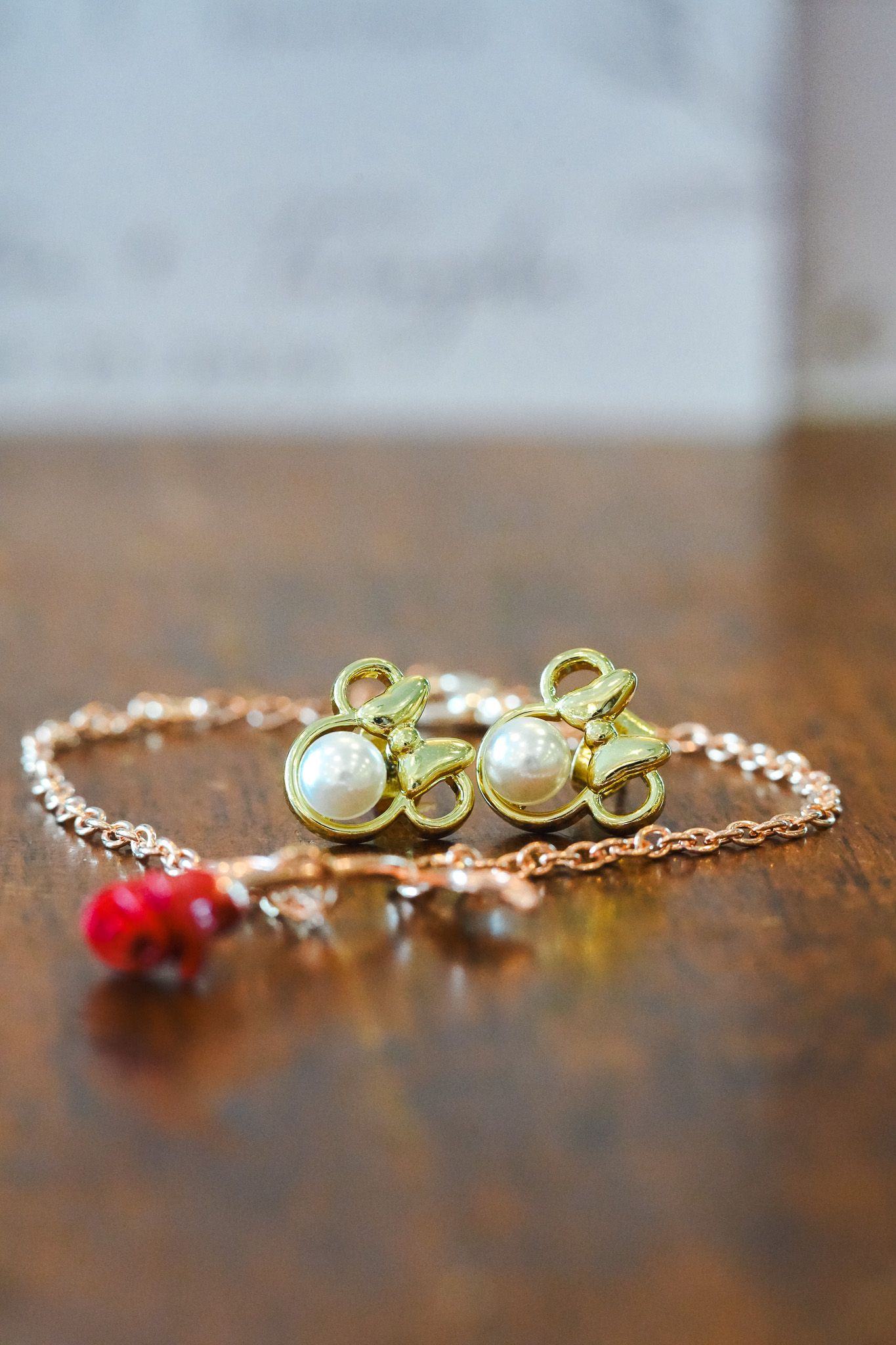 Gold pearl charm earrings beside a delicate rose gold chain bracelet with red coral bead on wooden surface.