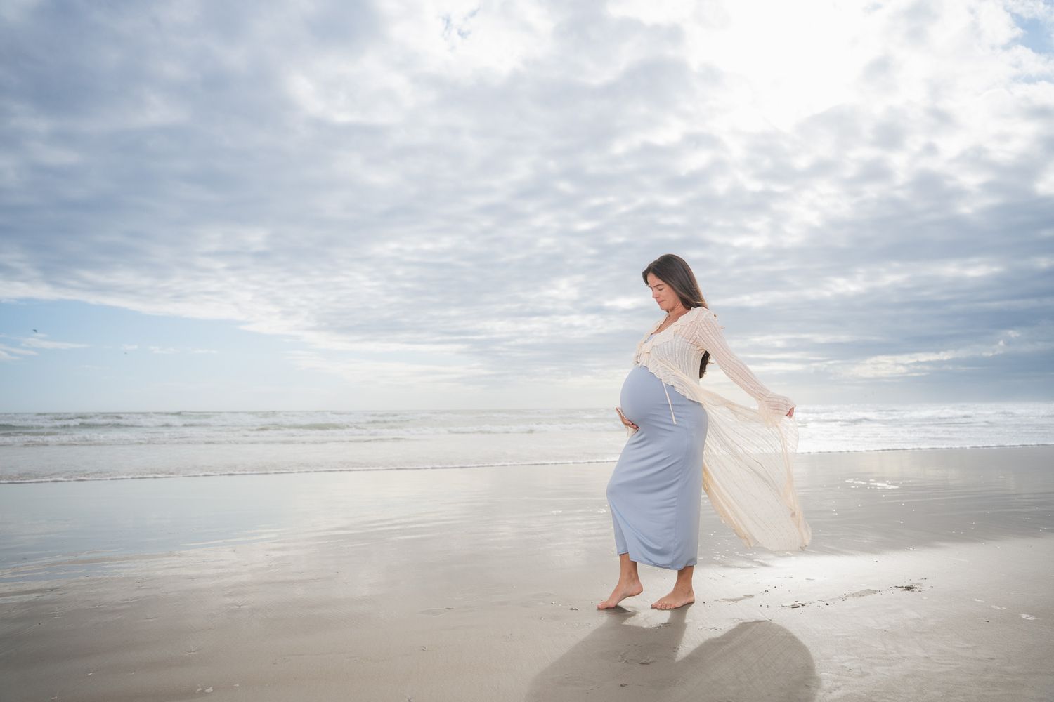 Pregnant woman in flowing blue dress and white wrap poses gracefully on a breezy beach at golden hour.