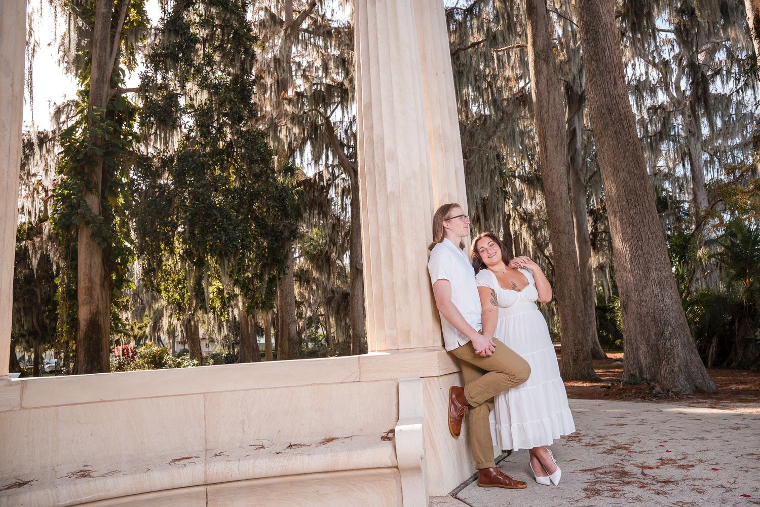 Couple holding hands and leaning against a white stone column in a sunlit park.