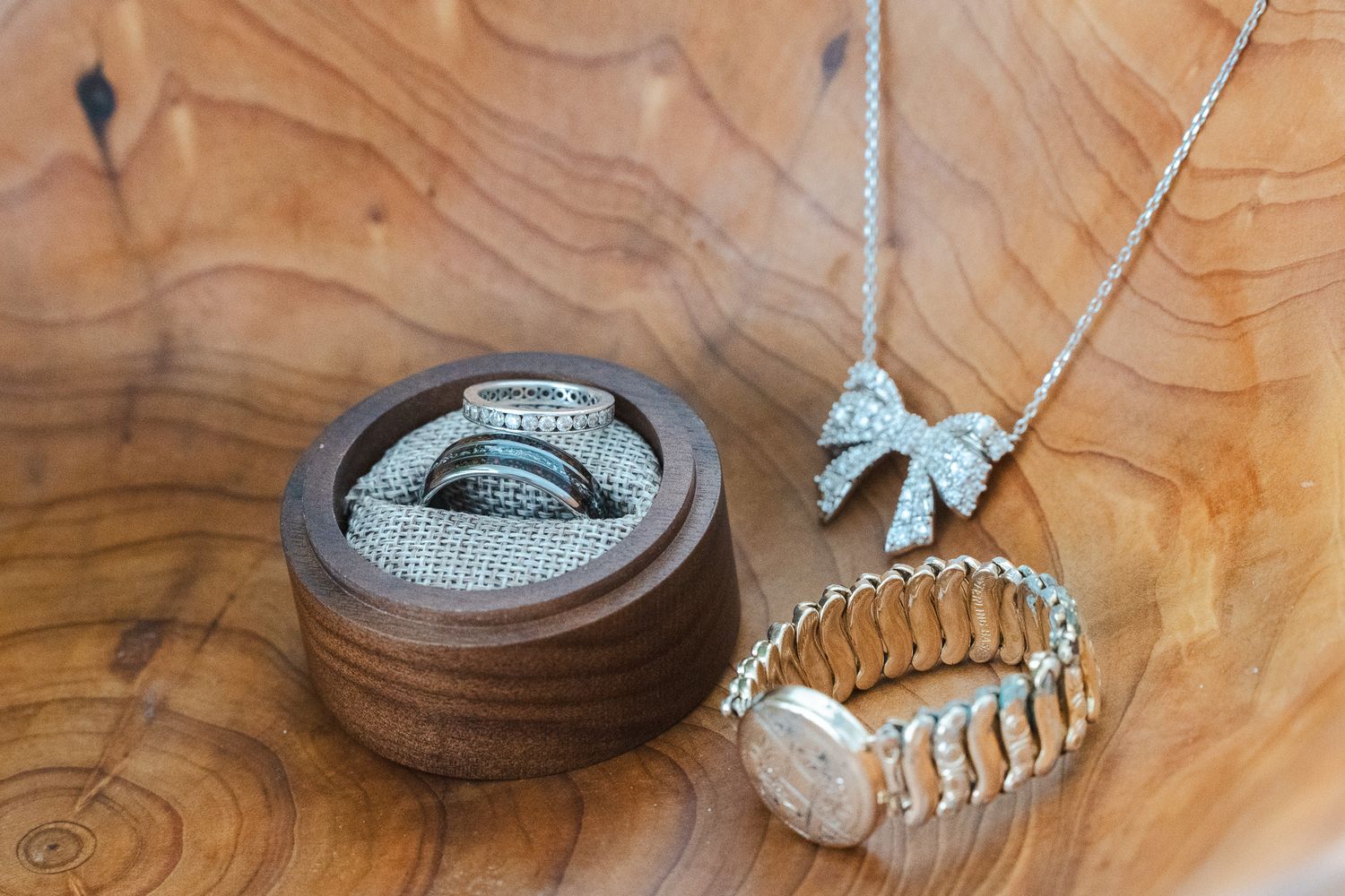 Wedding jewelry flat lay featuring diamond rings in wooden box, bow necklace, and gold bracelet.