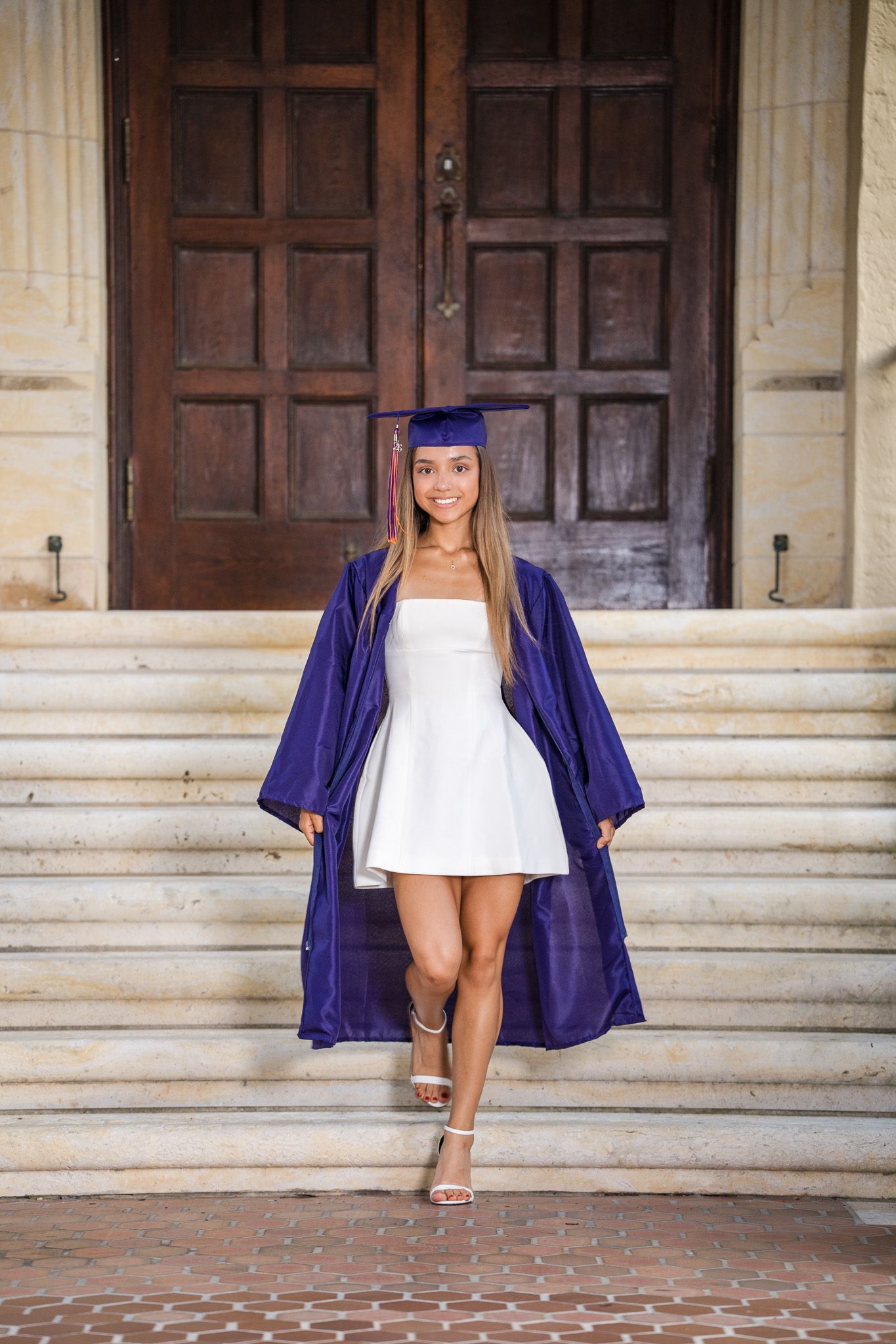 Smiling graduate in purple cap and gown with white dress posing on steps before large wooden doors.