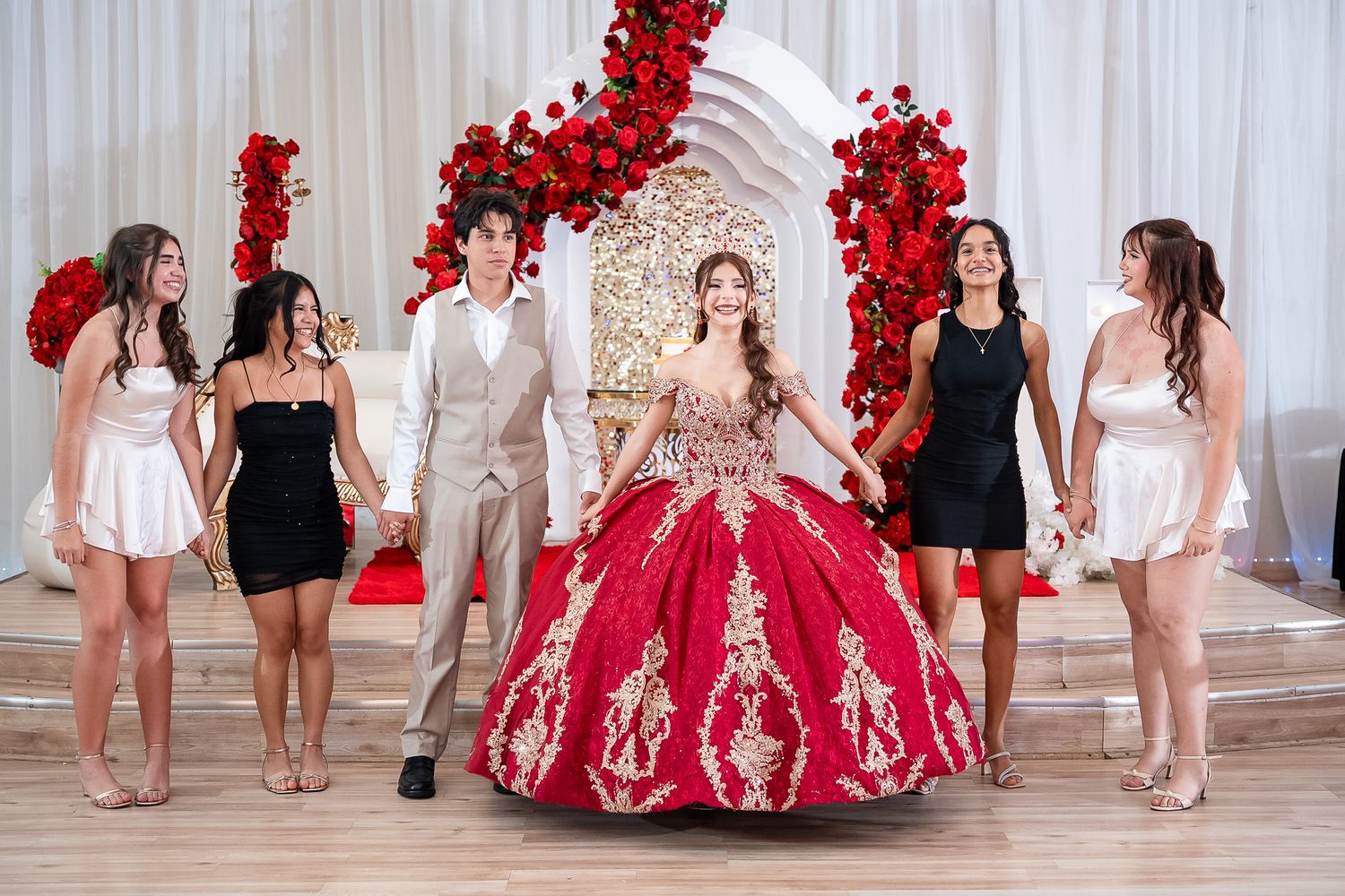 Quinceañera in red gold ballgown poses with court of honor before rose arch and throne backdrop.