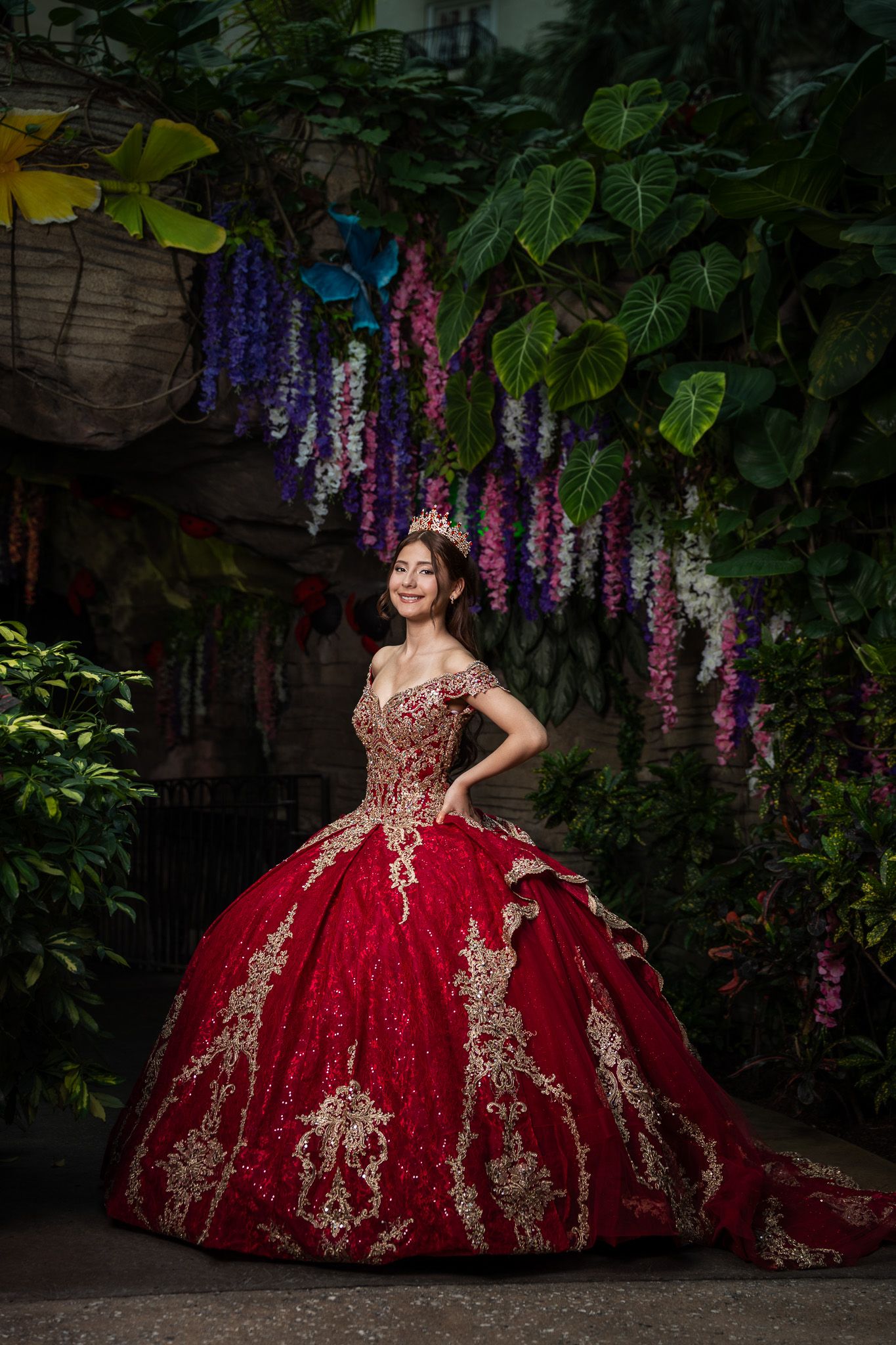 Young woman in red and gold quinceañera ball gown with tiara poses among lush greenery and wisteria flowers.