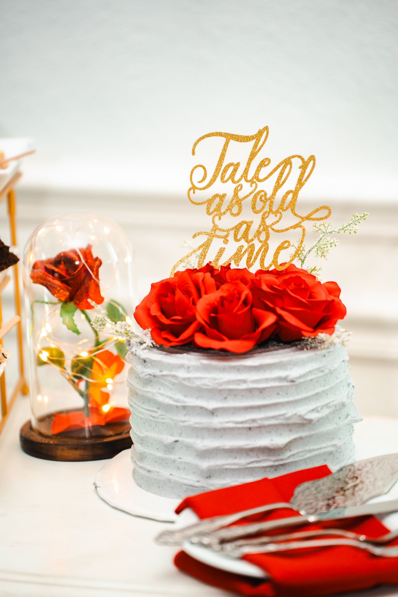 Elegant wedding cake with red roses and gold Tale as Old as Time topper on dessert table.