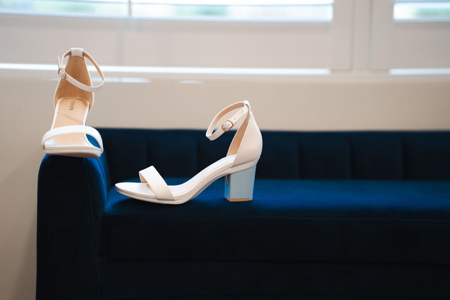 White block heel bridal sandals displayed on a navy blue velvet sofa.