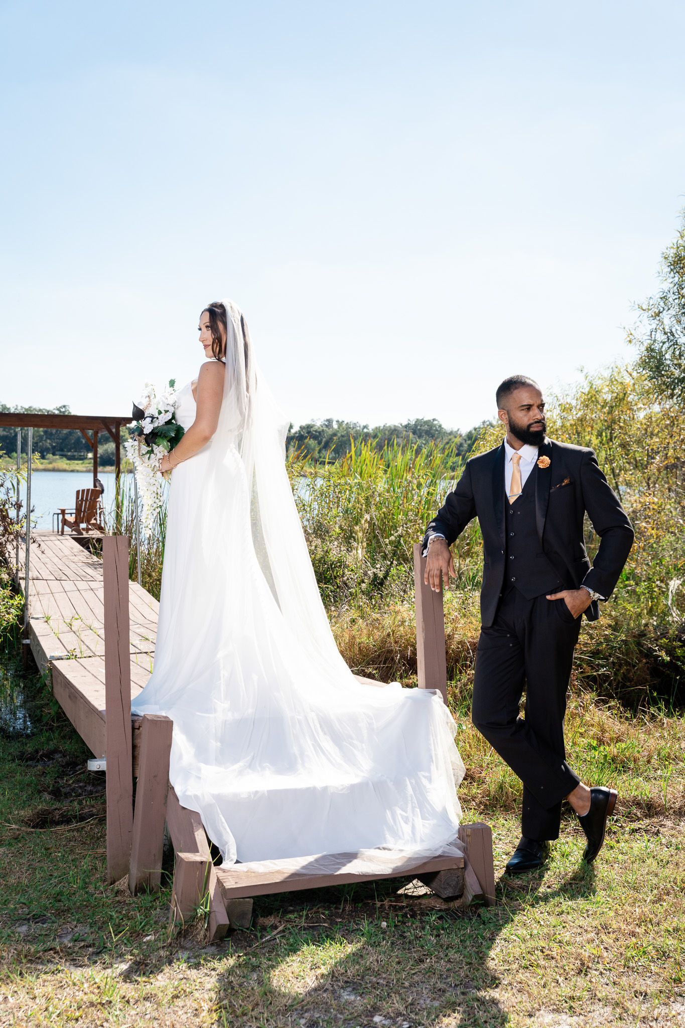 Bride in white gown with veil stands on dock steps while groom in black suit leans against railing outdoors.
