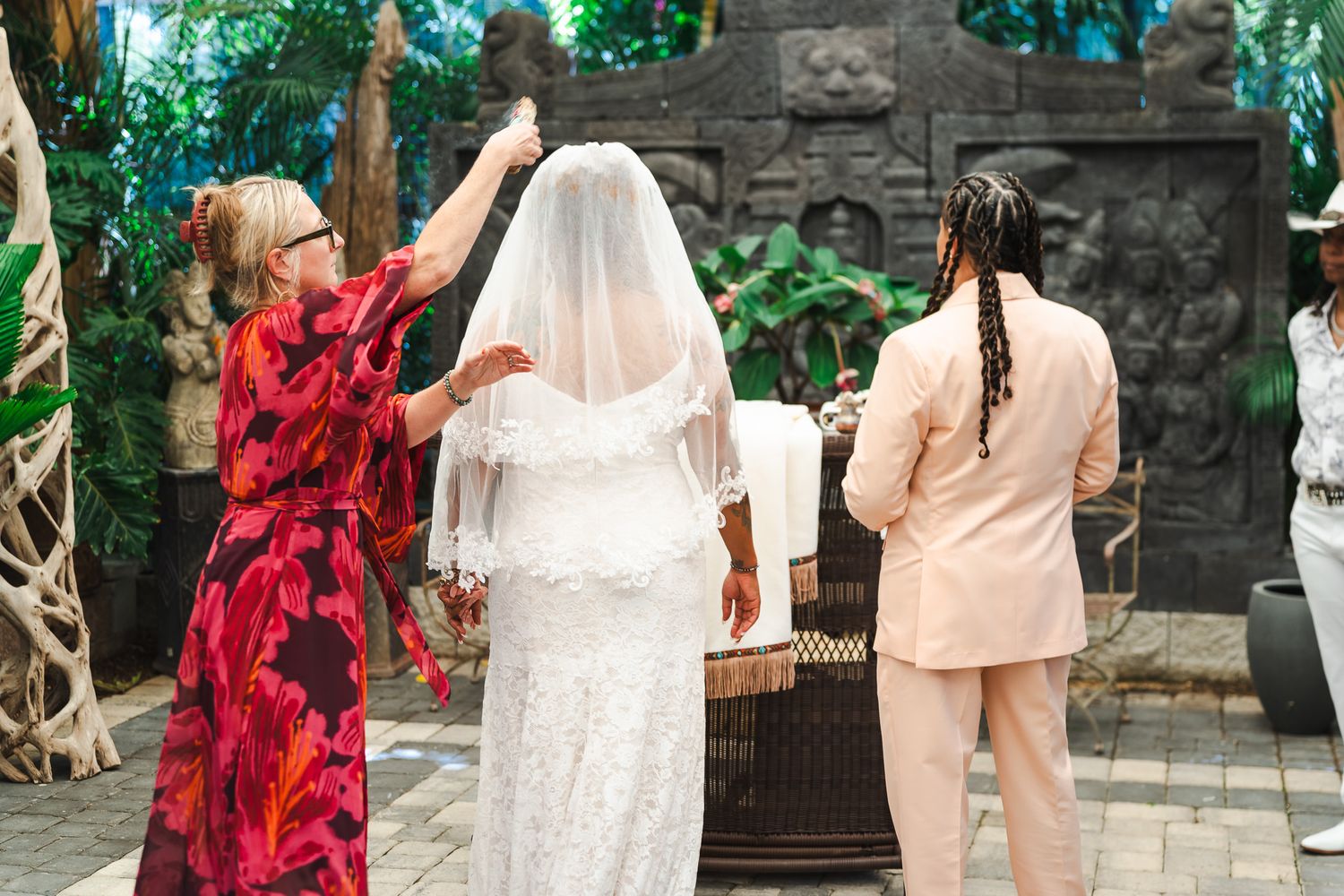Officiant in red floral dress lifts bride's veil during outdoor wedding ceremony.