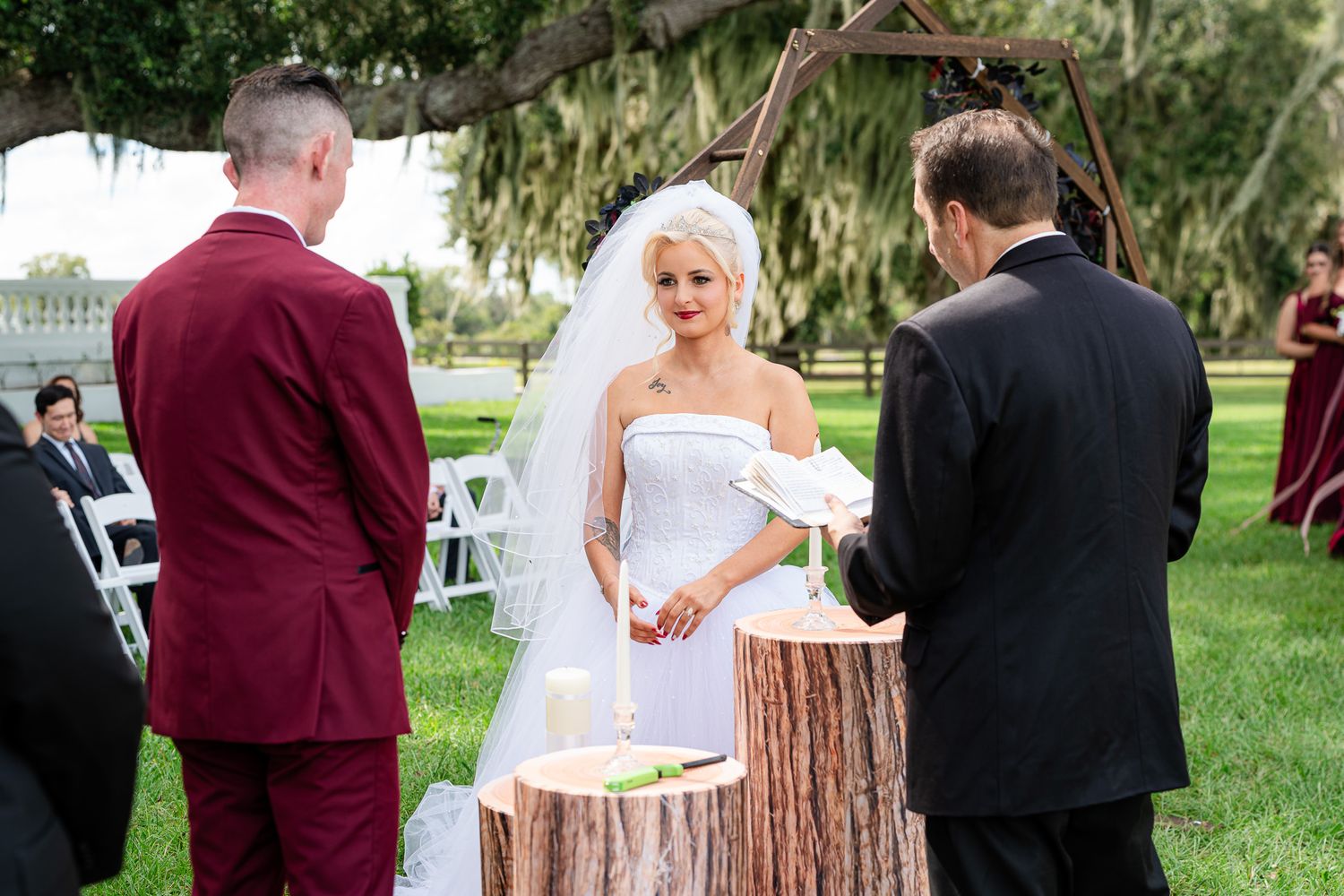 Blonde bride in strapless white gown exchanges vows at outdoor rustic wedding ceremony with wooden arch.