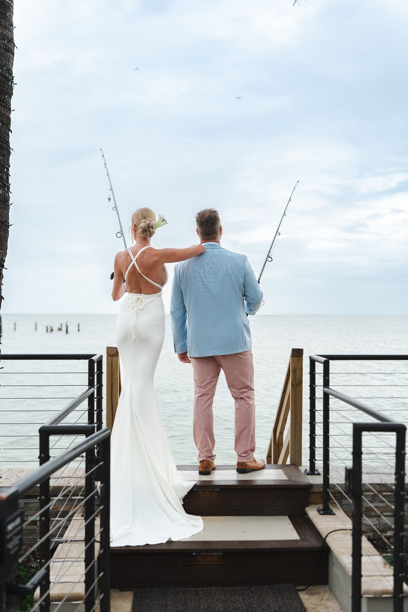 Bride and groom stand together holding fishing rods on waterfront dock with ocean view behind them.