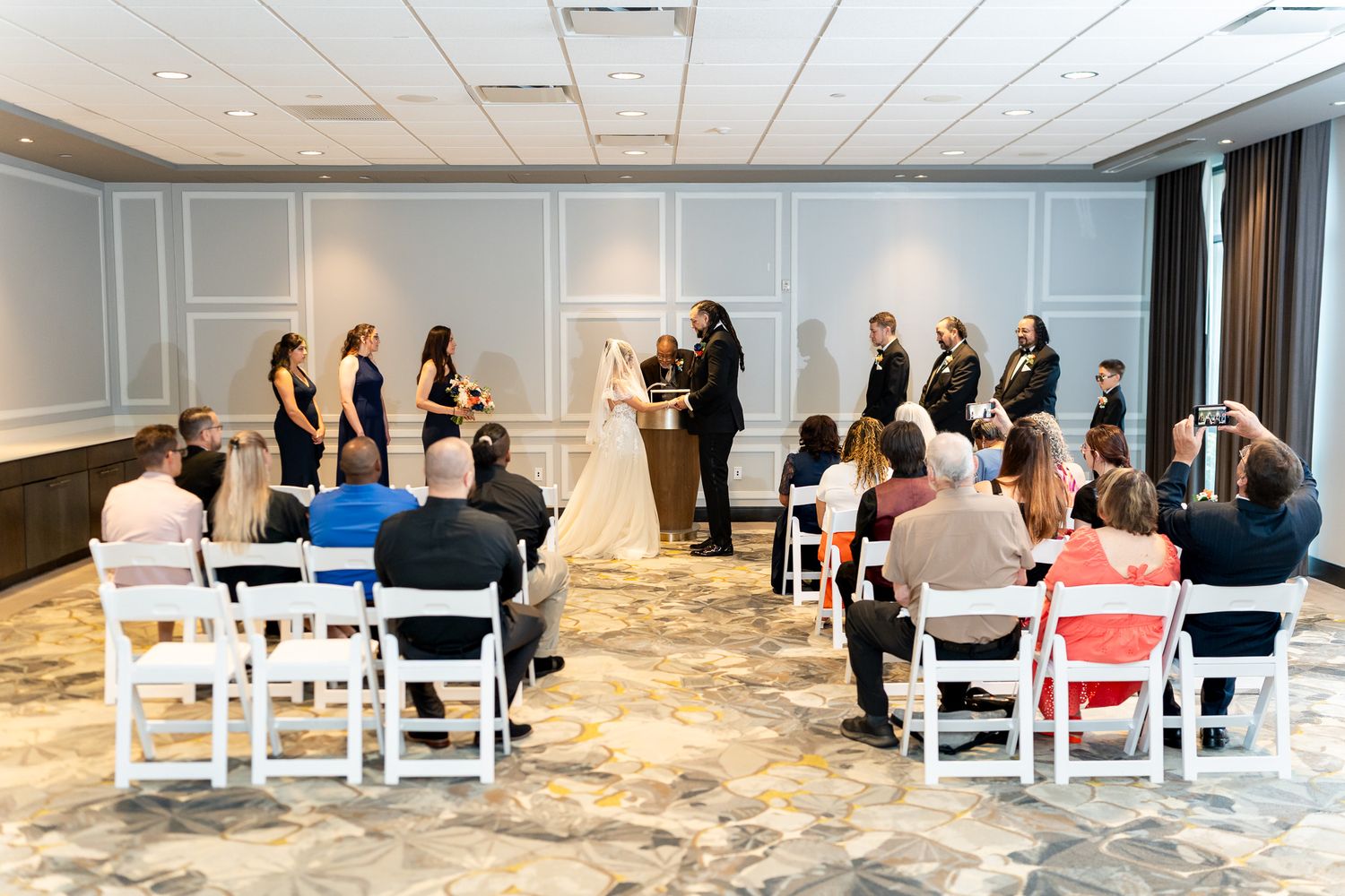 Wide shot of intimate indoor wedding ceremony with guests seated in white chairs around the couple.