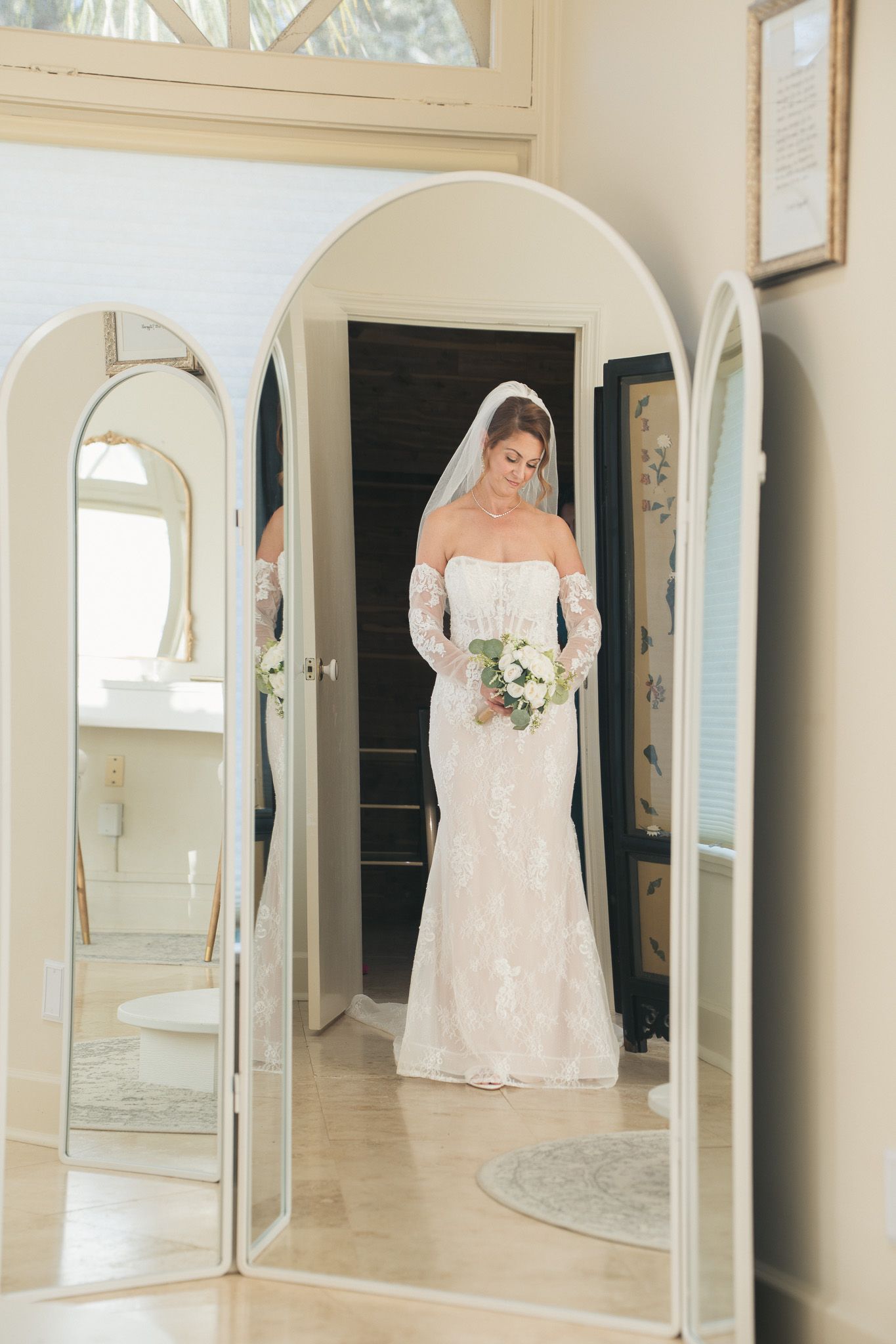 Bride in lace wedding gown holds bouquet while reflected in arched mirrors before ceremony.