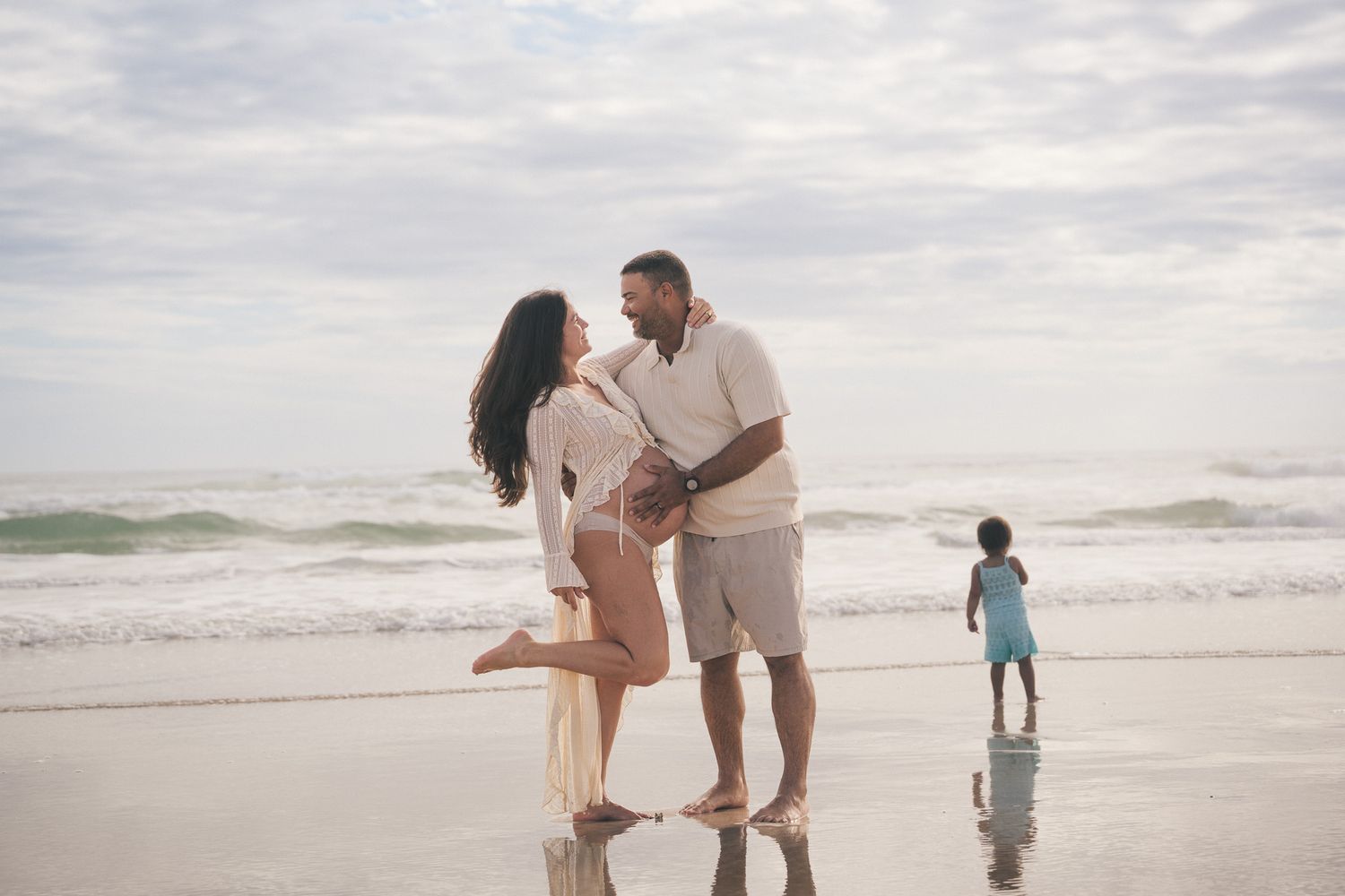 Couple sharing romantic moment on beach while child plays in ocean waves.