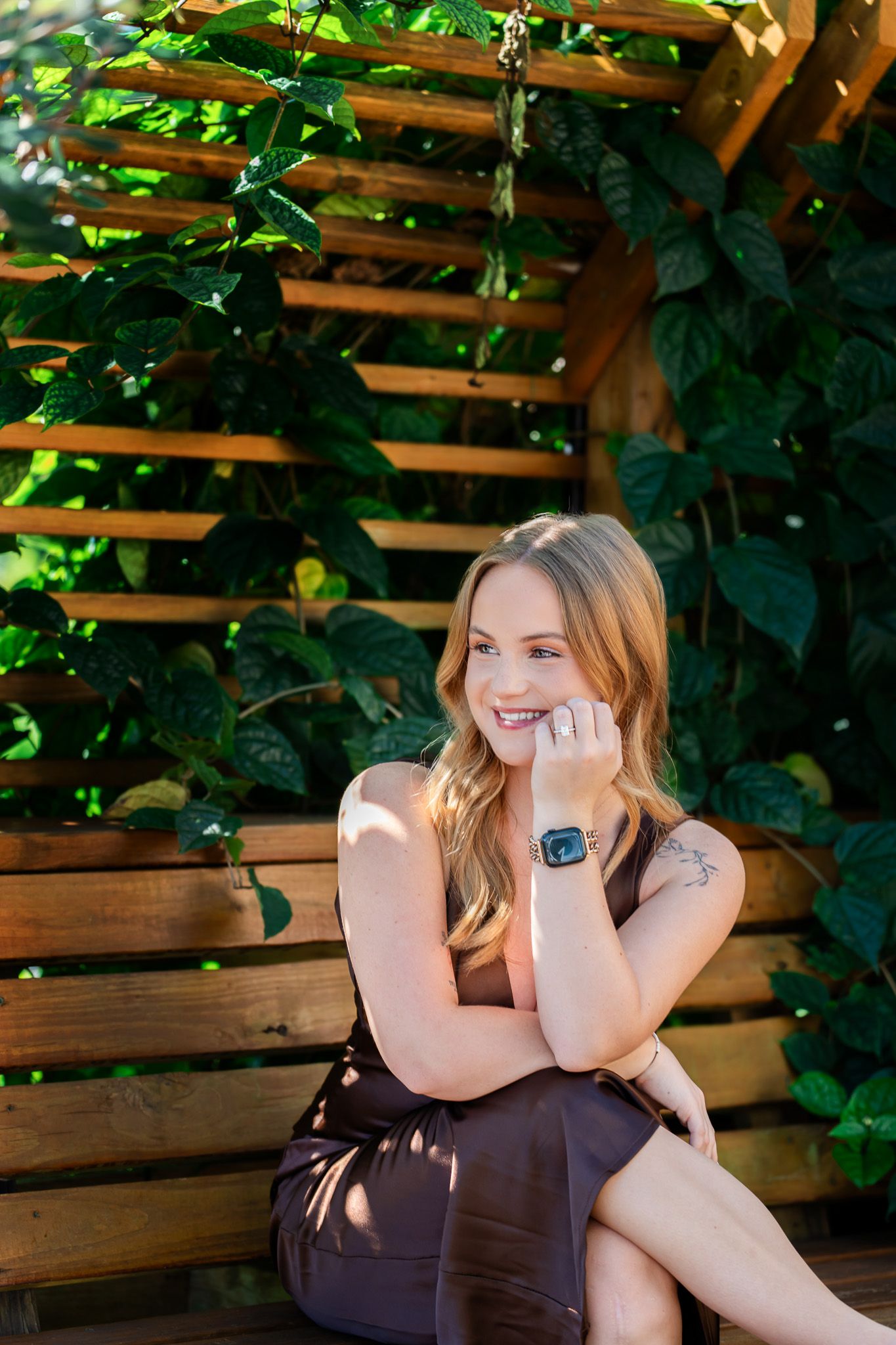 Smiling blonde woman in brown dress sitting on wooden bench surrounded by lush green foliage outdoors.