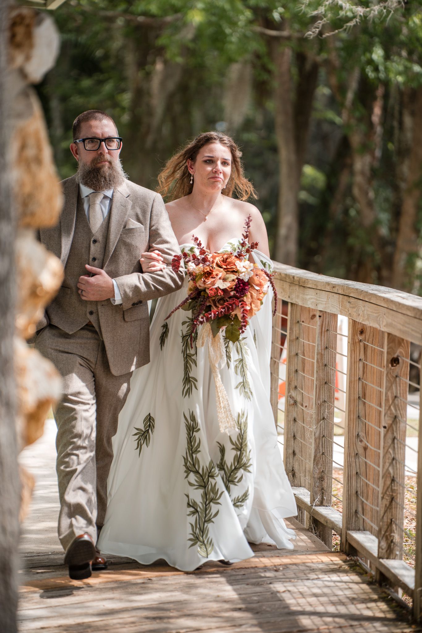 Bride in floral gown holding bouquet walks arm-in-arm with bearded groom in tan suit on wooden bridge.