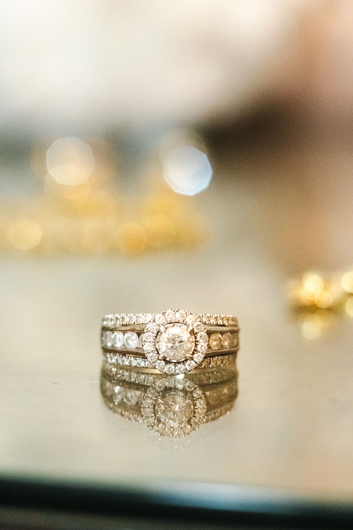 Diamond halo engagement ring stacked with wedding bands reflected on a glossy surface with bokeh background.