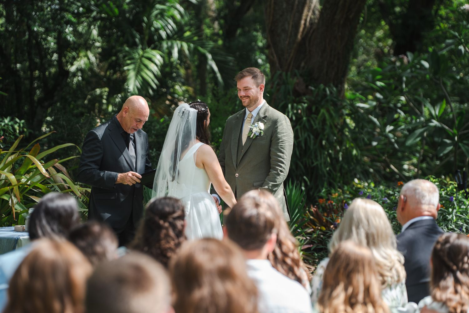 Bride and groom hold hands during outdoor wedding ceremony surrounded by lush tropical greenery.