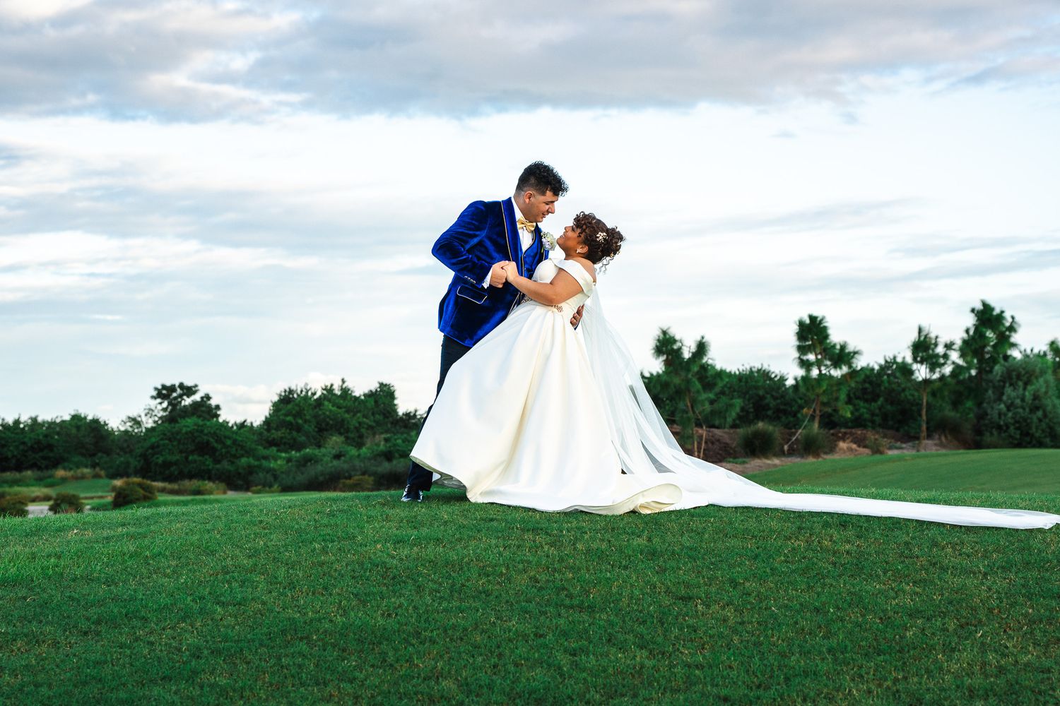 Groom dips bride in white ballgown on lush green lawn during romantic outdoor wedding photo session.