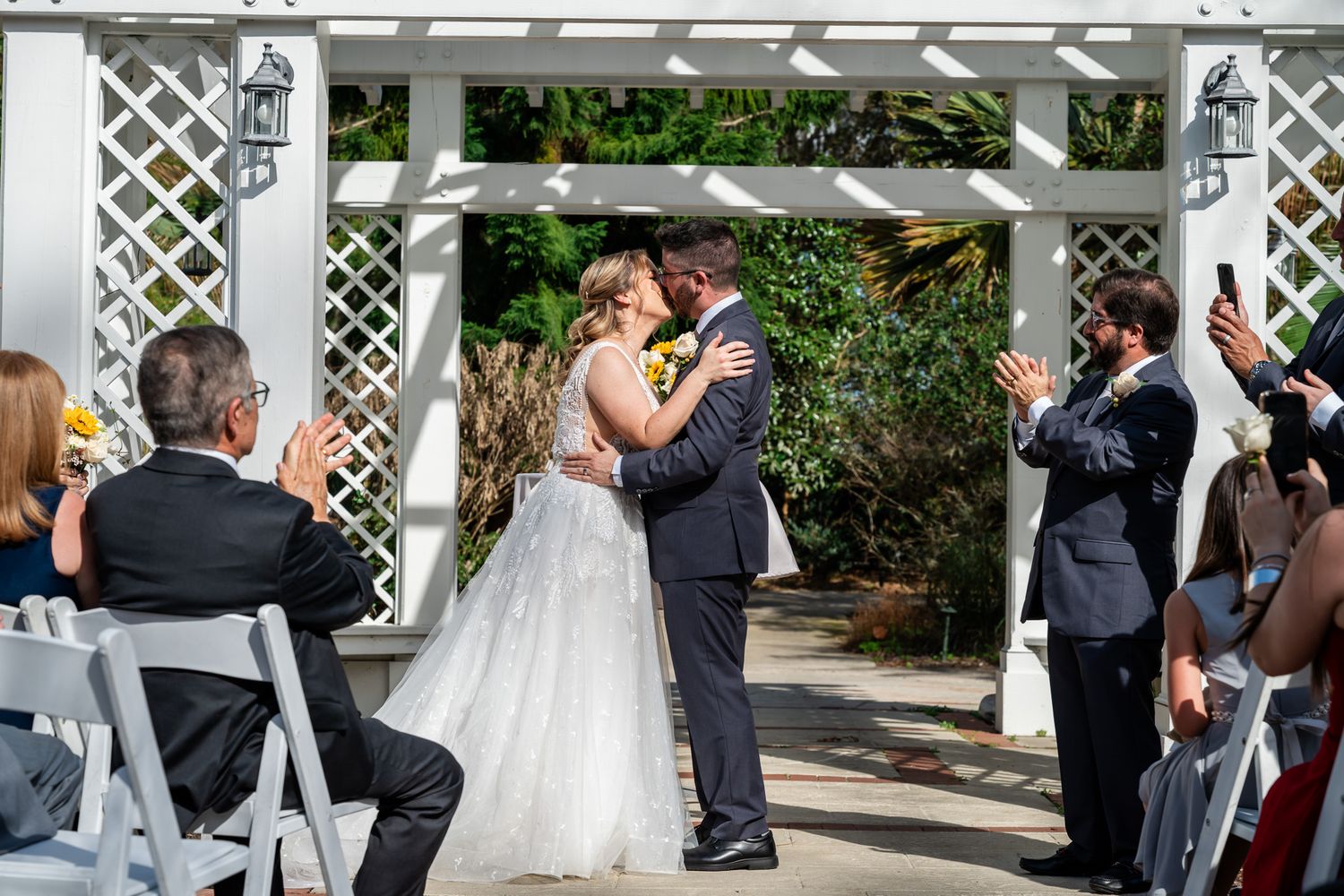 Bride and groom share first kiss under white lattice pergola as guests applaud and capture the moment on phones.