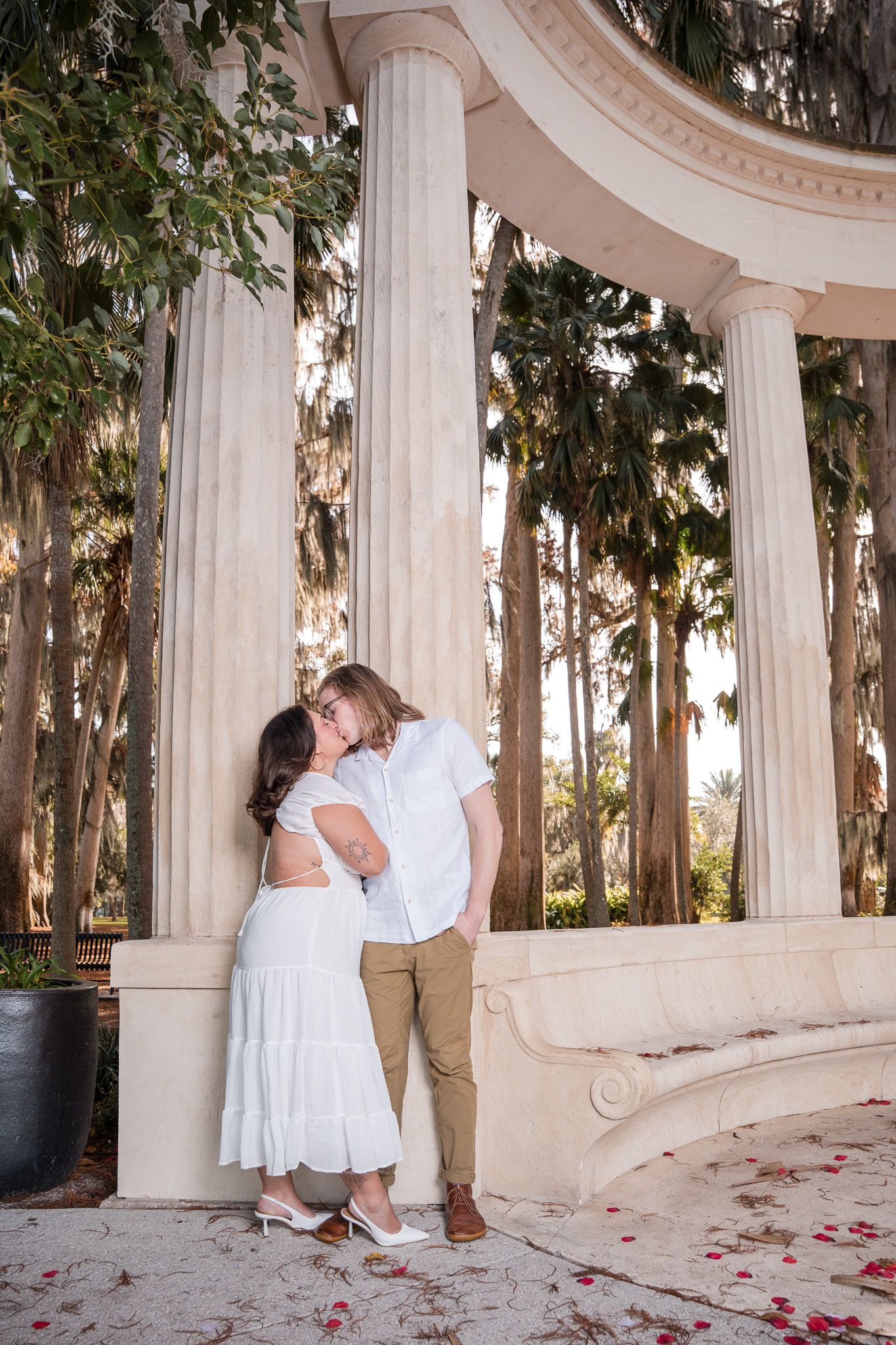 Couple sharing a kiss beside tall white columns with rose petals on the ground.