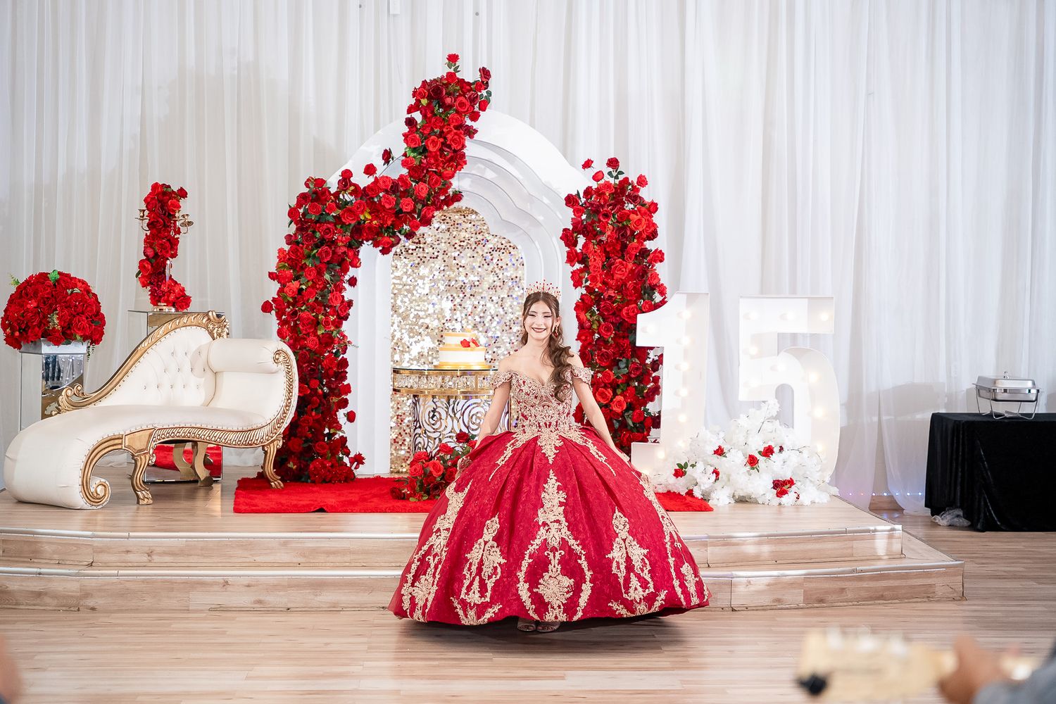Quinceañera girl in red gold gown stands on stage with rose arch and throne chair decor.