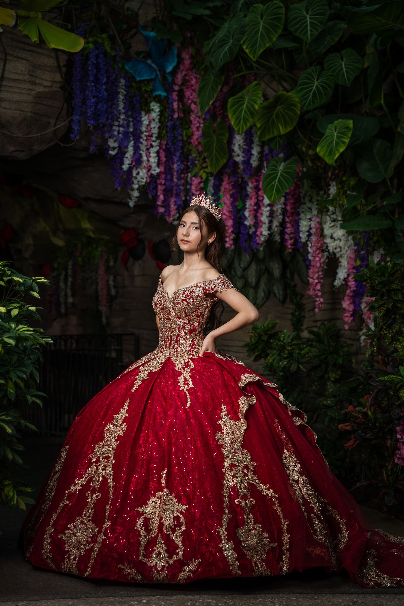 Young woman in red and gold ball gown with crown poses among hanging wisteria flowers in lush garden setting.