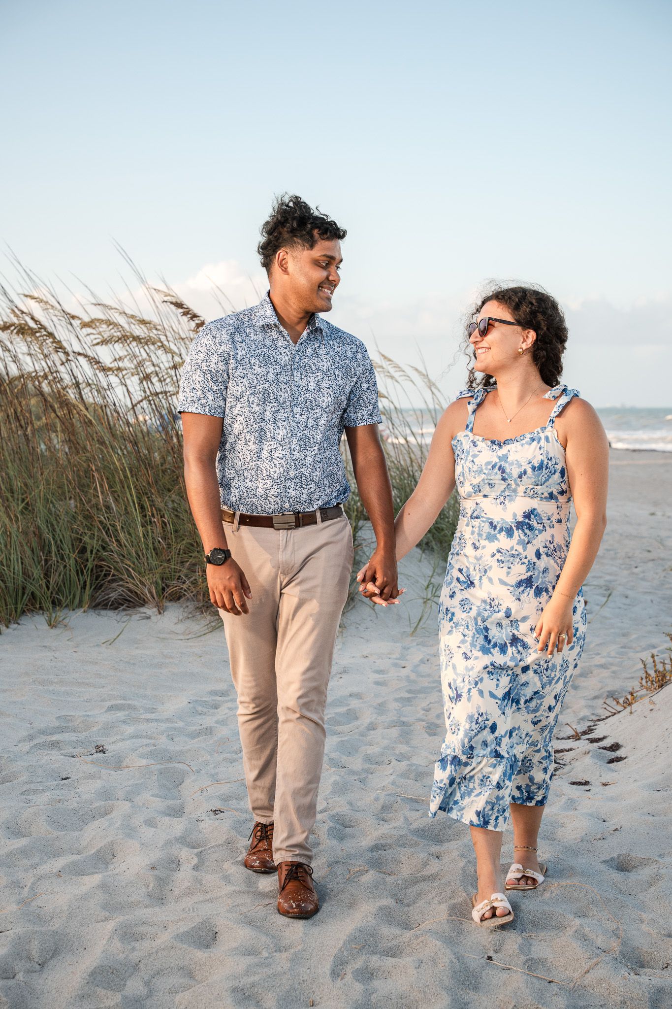Couple holding hands walking on sandy beach, man in blue floral shirt and woman in blue floral dress.
