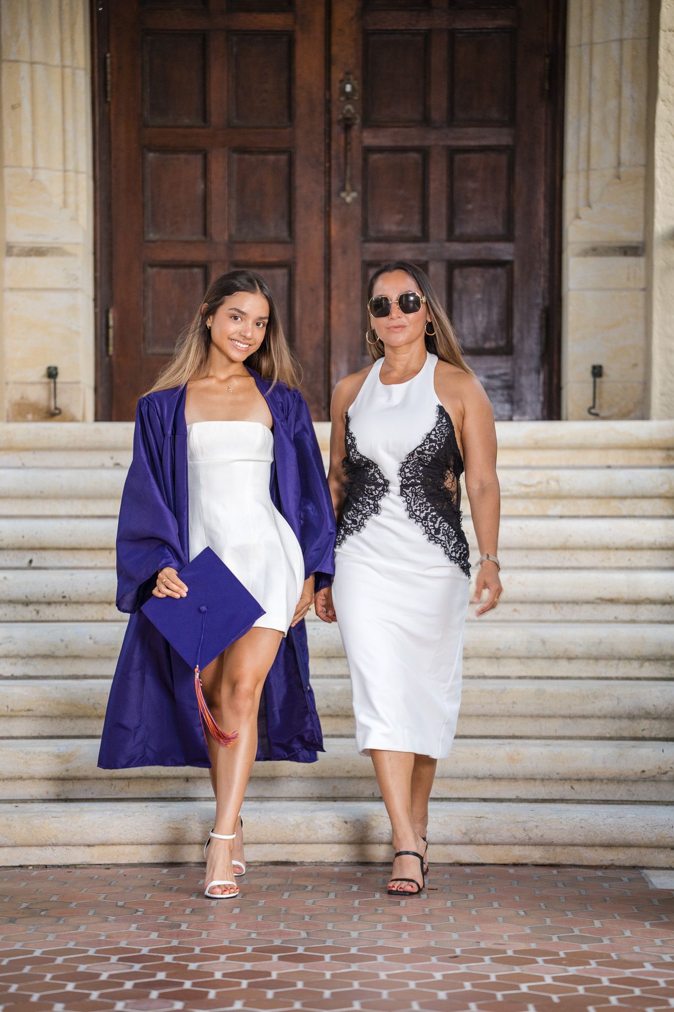 A smiling graduate in purple robe holds cap while walking with woman in white dress on university steps.