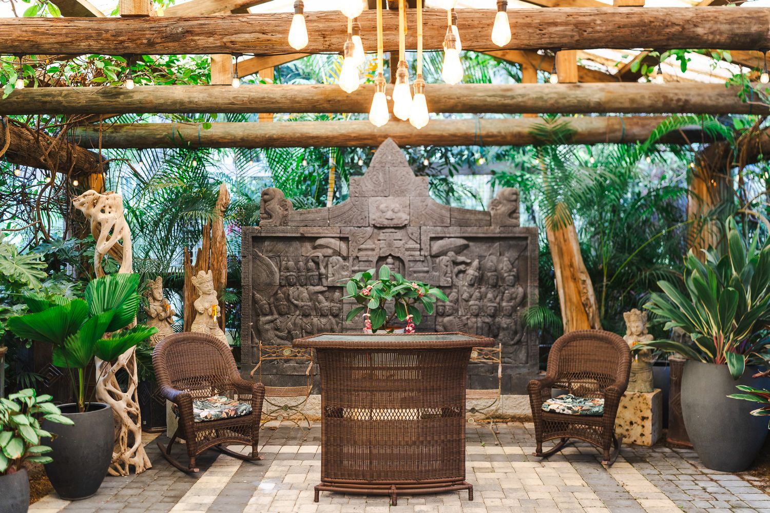 Tropical garden patio with wicker furniture, hanging Edison bulbs, and an ornate stone Mayan relief wall.